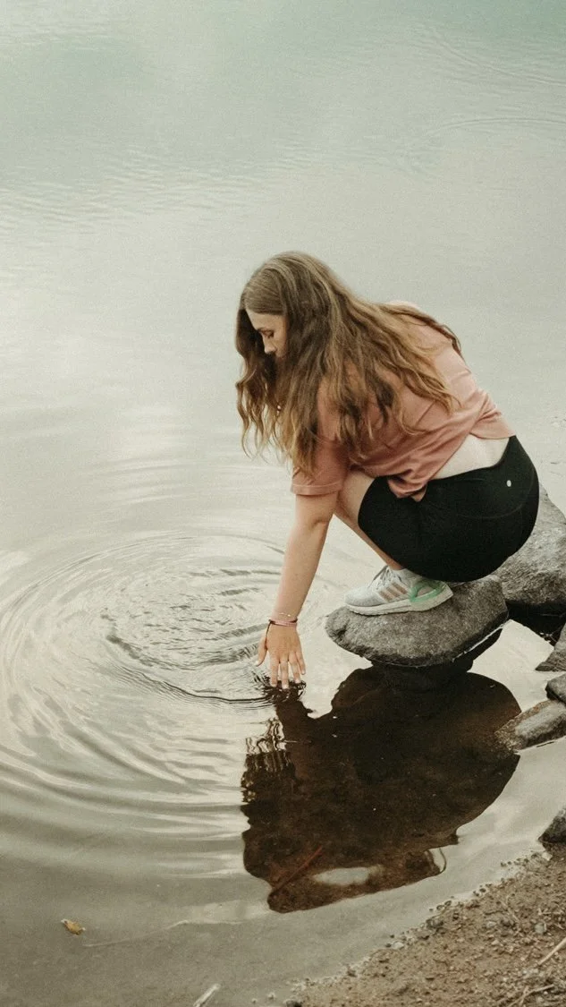 A young woman with long brown hair crouching on rocks by a calm lake, dipping her hand into the water.