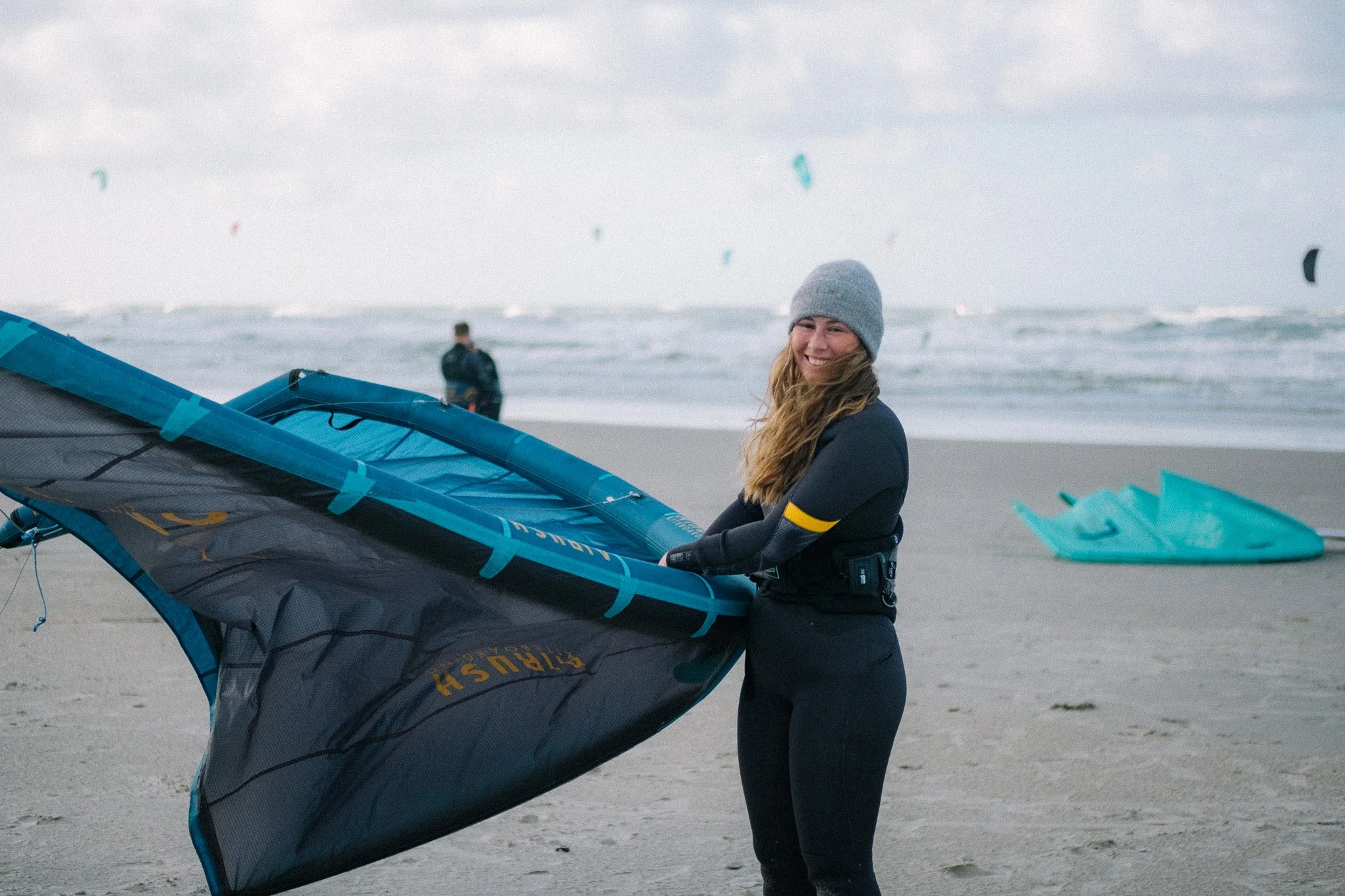 A woman in a gray beanie and black wetsuit smiling while holding a blue windsurfing sail on a beach with water, sand, and other windsurfing sails visible in the background.
