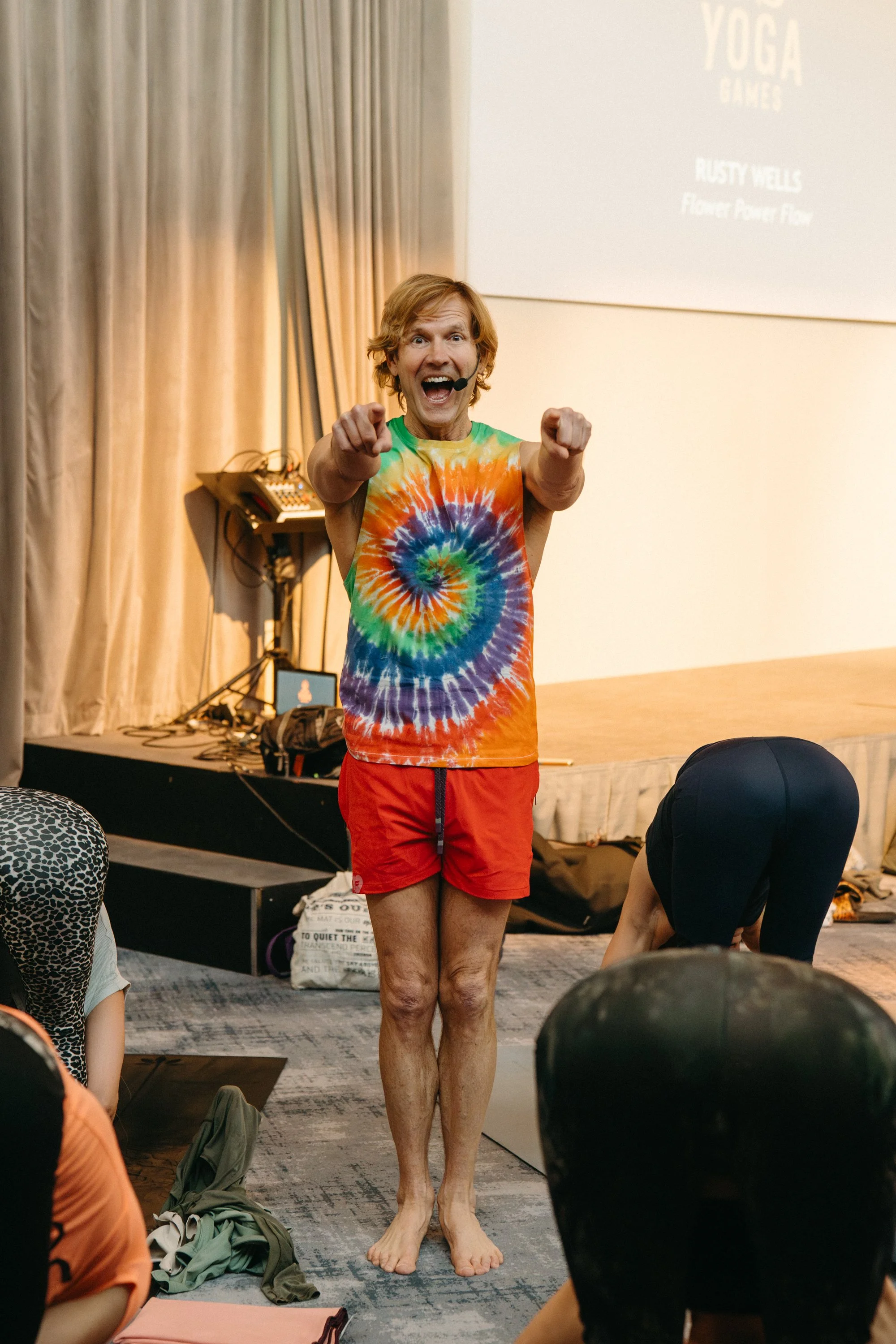 An instructor in a colorful tie-dye shirt and red shorts leading a yoga class, standing on a mat and pointing towards the camera with an energetic expression.