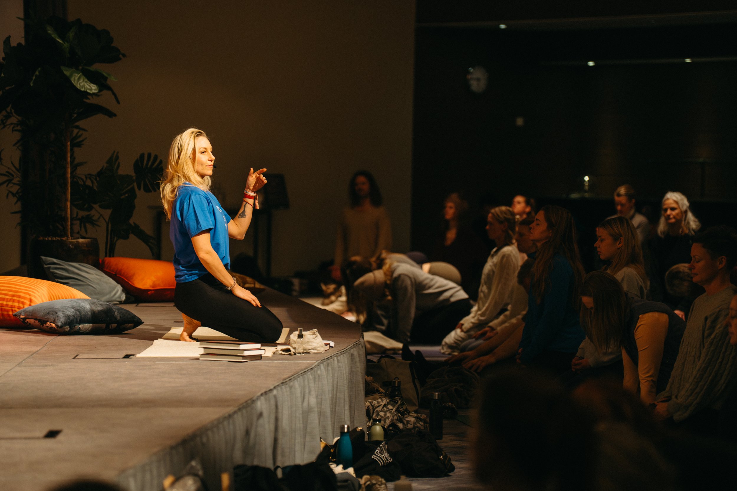 A woman in a blue shirt kneels on stage and gestures while speaking to an audience seated on the floor in a dark room, with pillows and personal items around.