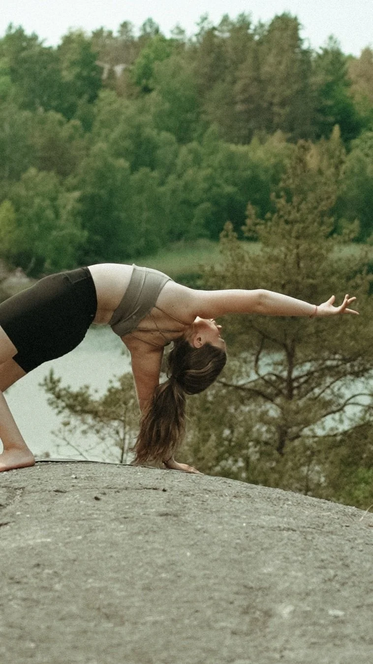 A woman practicing yoga outdoors on a large rock, performing a backbend with one arm extended forward, surrounded by trees and water.