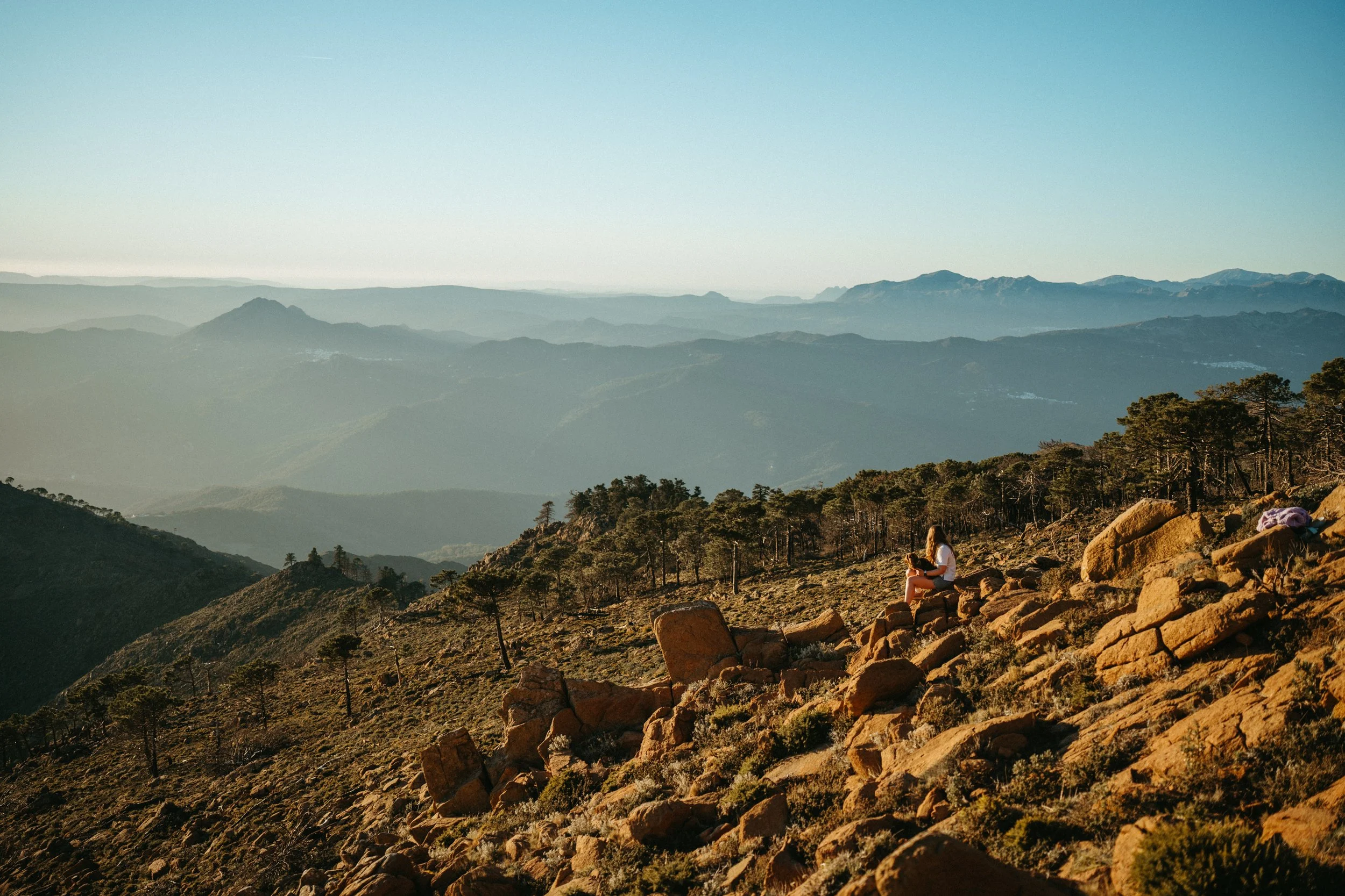 A person sitting on rocks on a mountain slope, overlooking a vast range of mountains with trees and a clear sky.