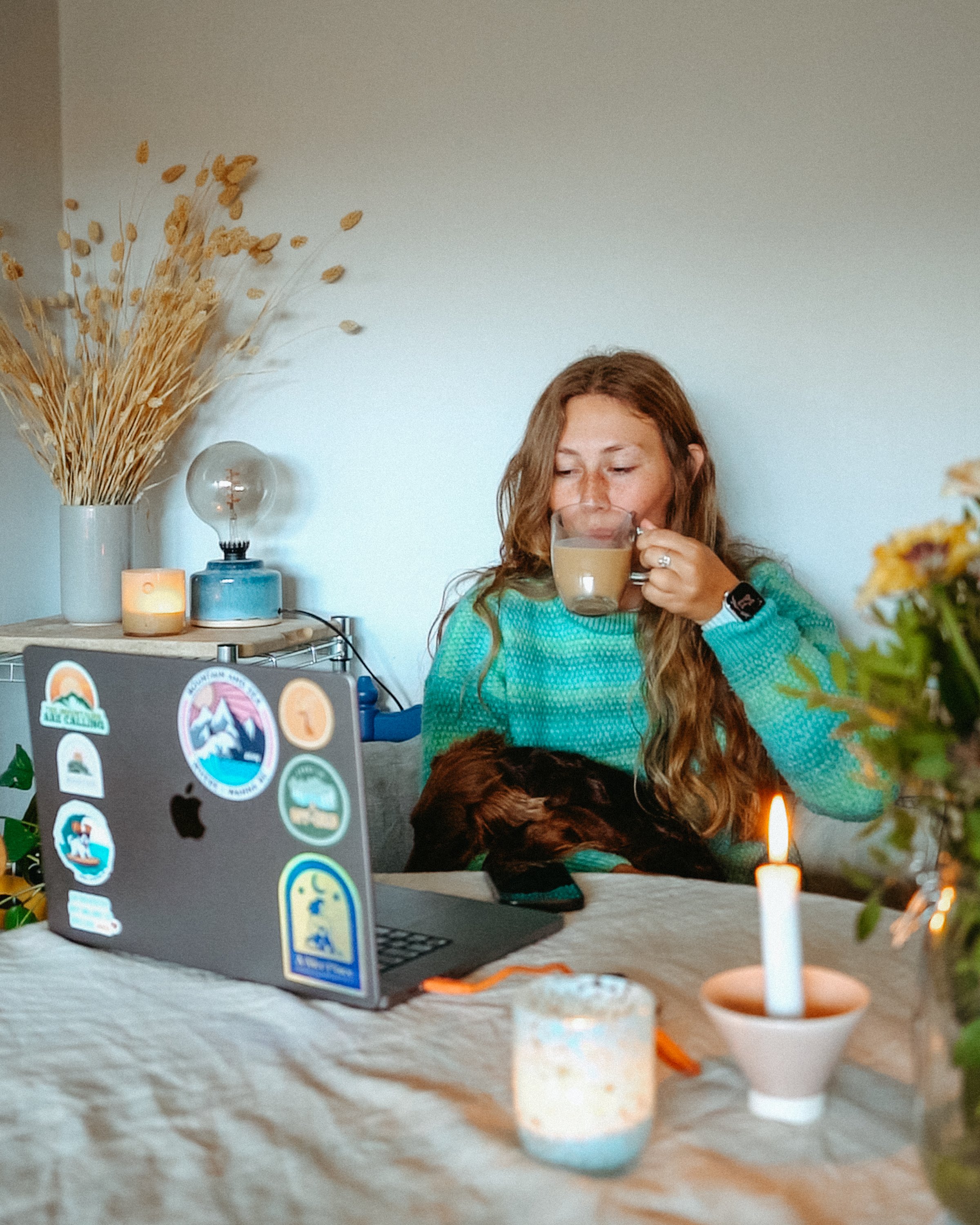 A woman sitting at a table drinking coffee with a laptop open in front of her, decorated with stickers, in a cozy room with candles and dried flowers.