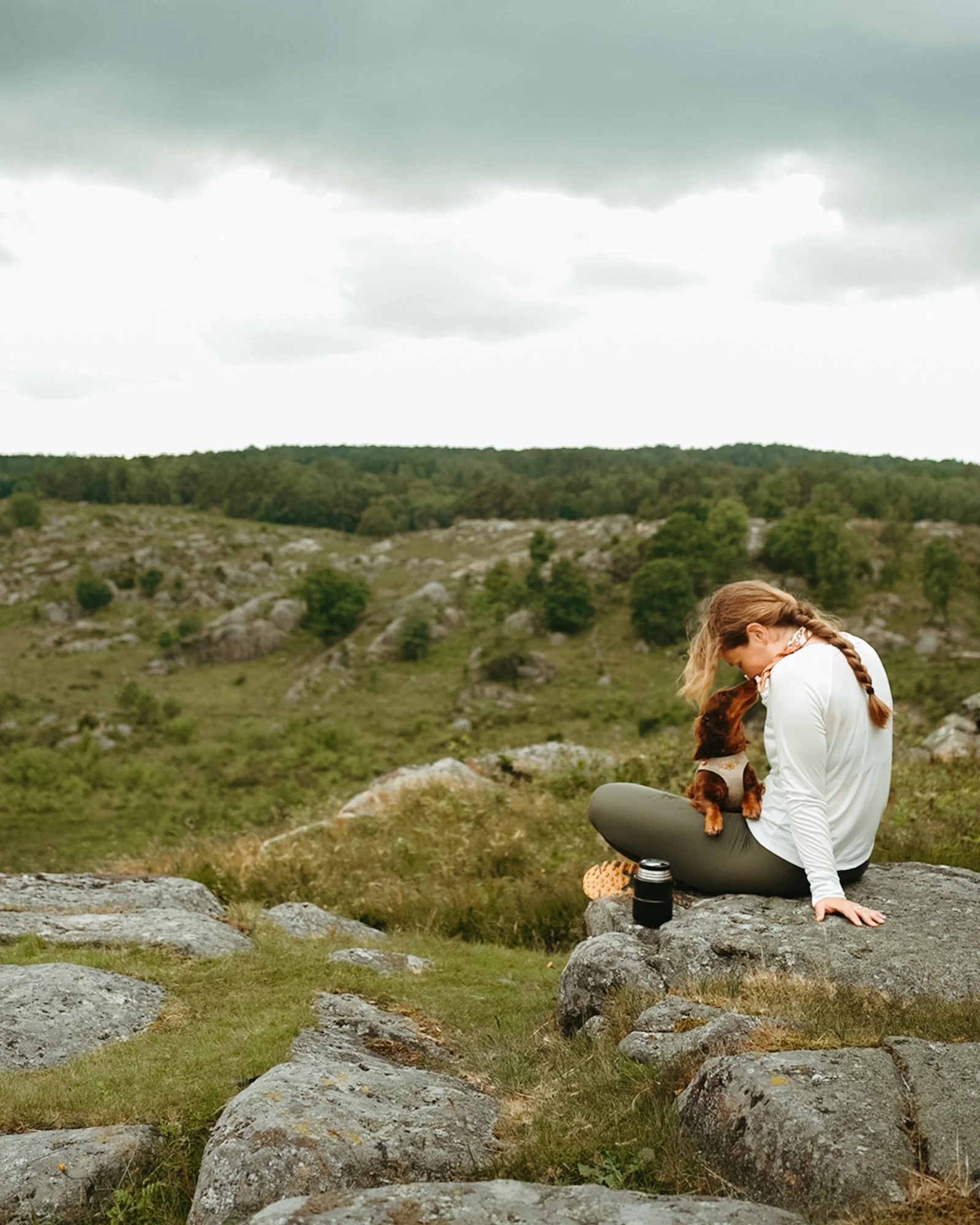 Woman sitting on a rock outdoors with a small dog, in a grassy and rocky landscape under a cloudy sky.
