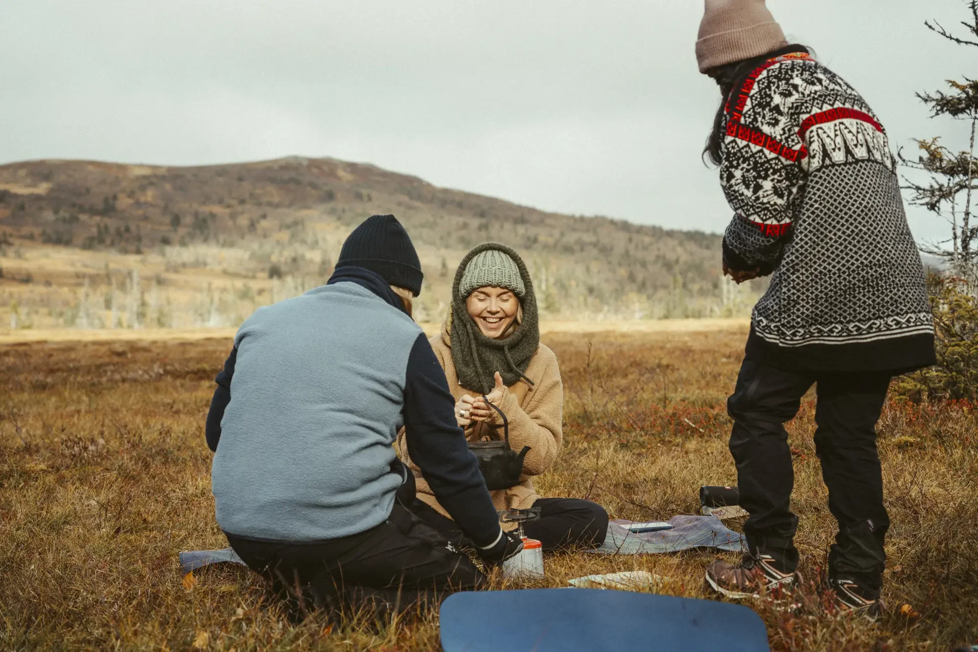 Three friends outdoors in a grassy field during fall, sitting and standing, wearing warm clothing, smiling, with mountains in the background.