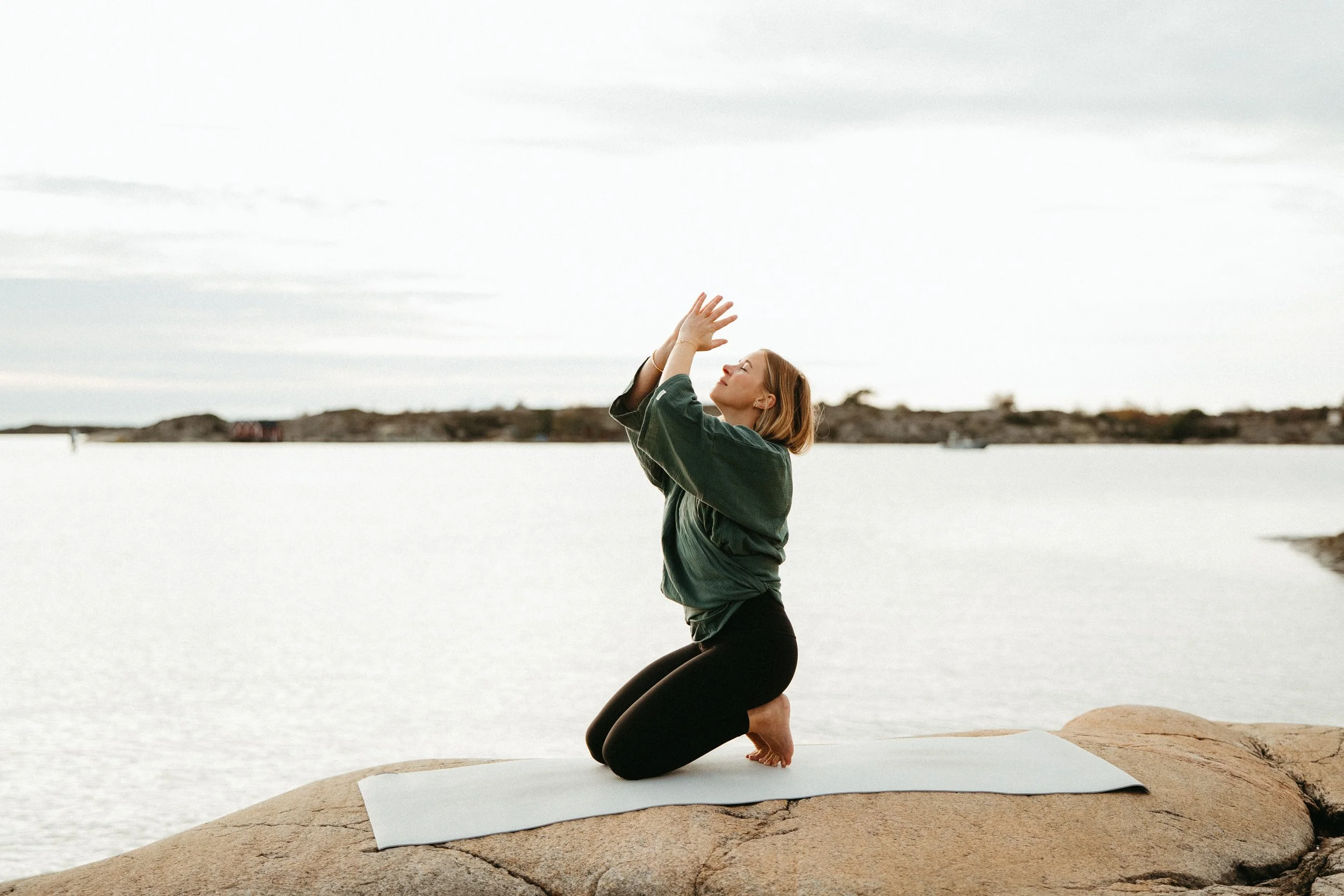 A woman practicing yoga outdoors on a rock near a body of water, kneeling on a yoga mat, with her hands raised above her head and eyes closed.
