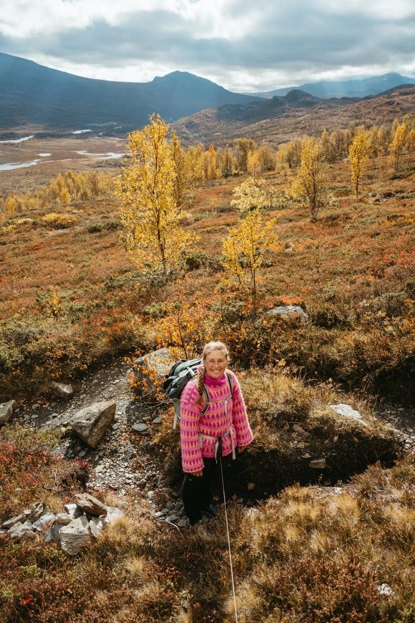 A woman hiking in a mountainous landscape with fall foliage, wearing a pink patterned sweater and carrying a backpack, smiling at the camera.