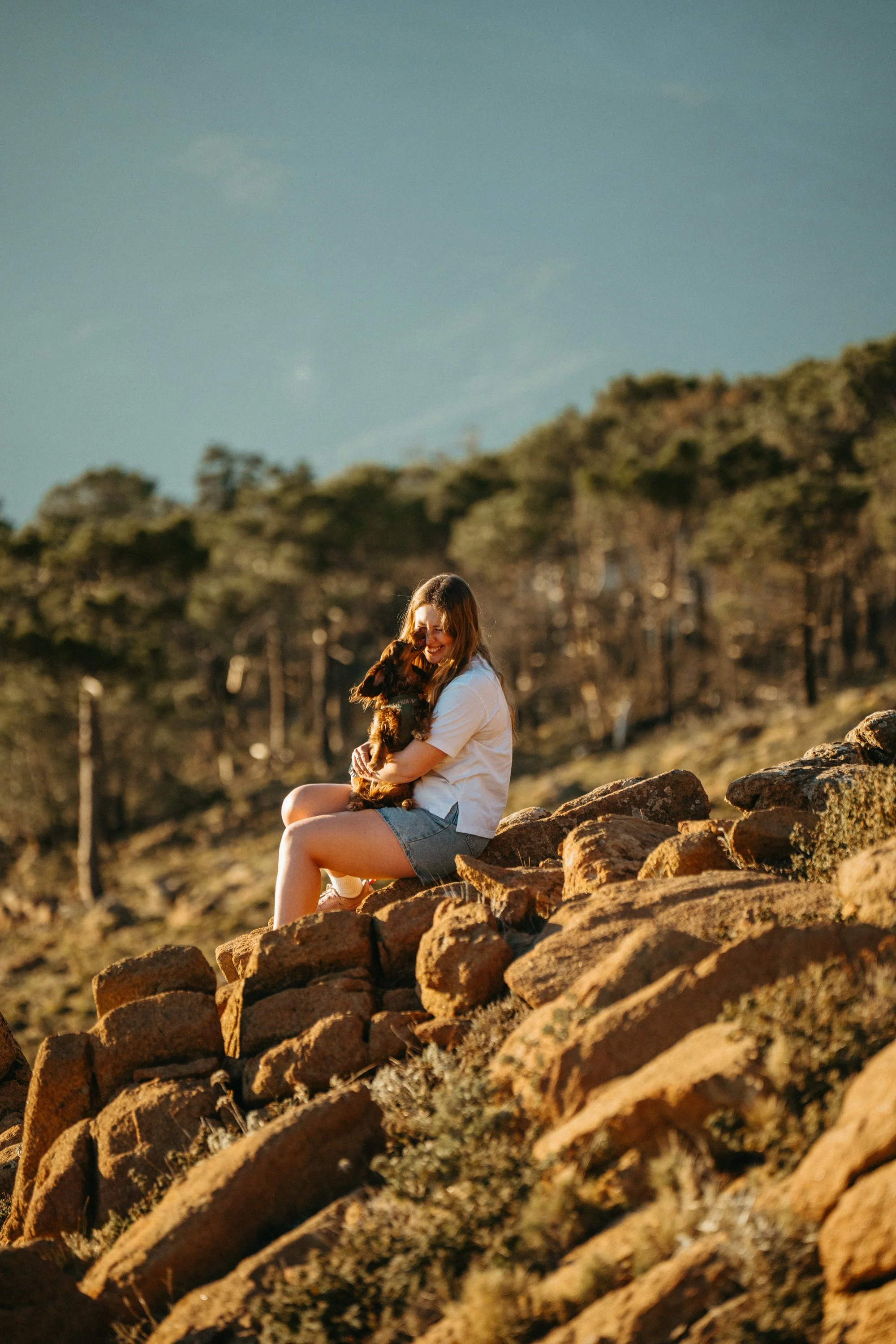 A woman sitting on rocks outdoors, holding a dog in her arms, smiling at the dog, with a forest and blue sky in the background during sunset.