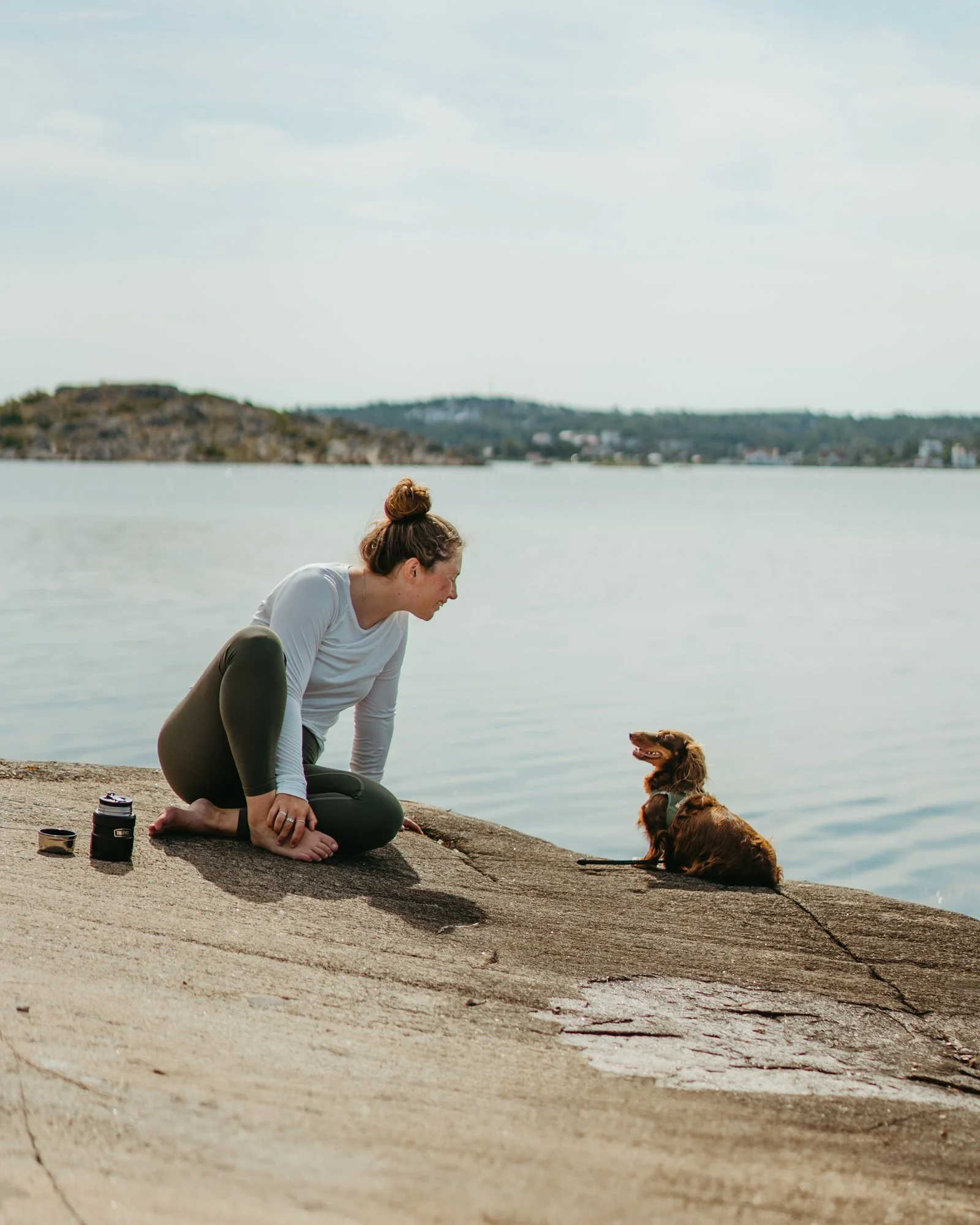 A woman and a dog sitting on a concrete surface near a body of water, with distant hills in the background, during daytime.