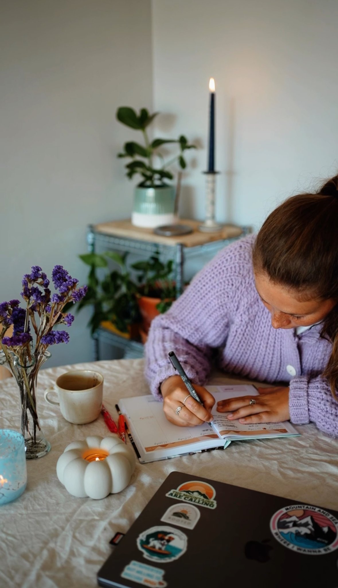 A woman in a lavender sweater writing in a planner on a table, with a white pumpkin candle, a cup of tea, and a vase of purple flowers in the foreground. In the background, there is a small table with potted plants, a candle holder, and a lit candle.