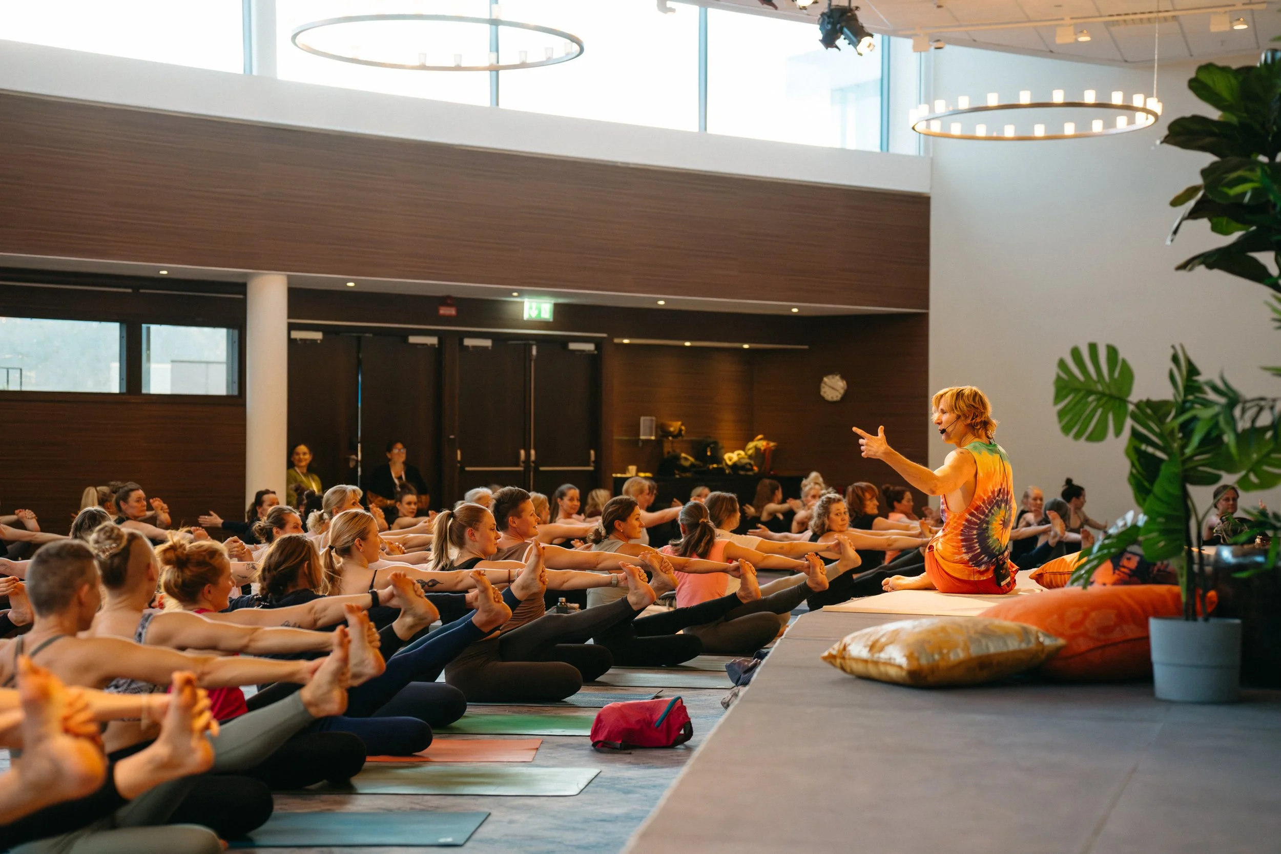 A large group of women participating in a yoga class or workshop, sitting cross-legged and stretching their arms forward, led by an instructor sitting on a raised platform in a modern, bright indoor space with large windows and decorative lighting.