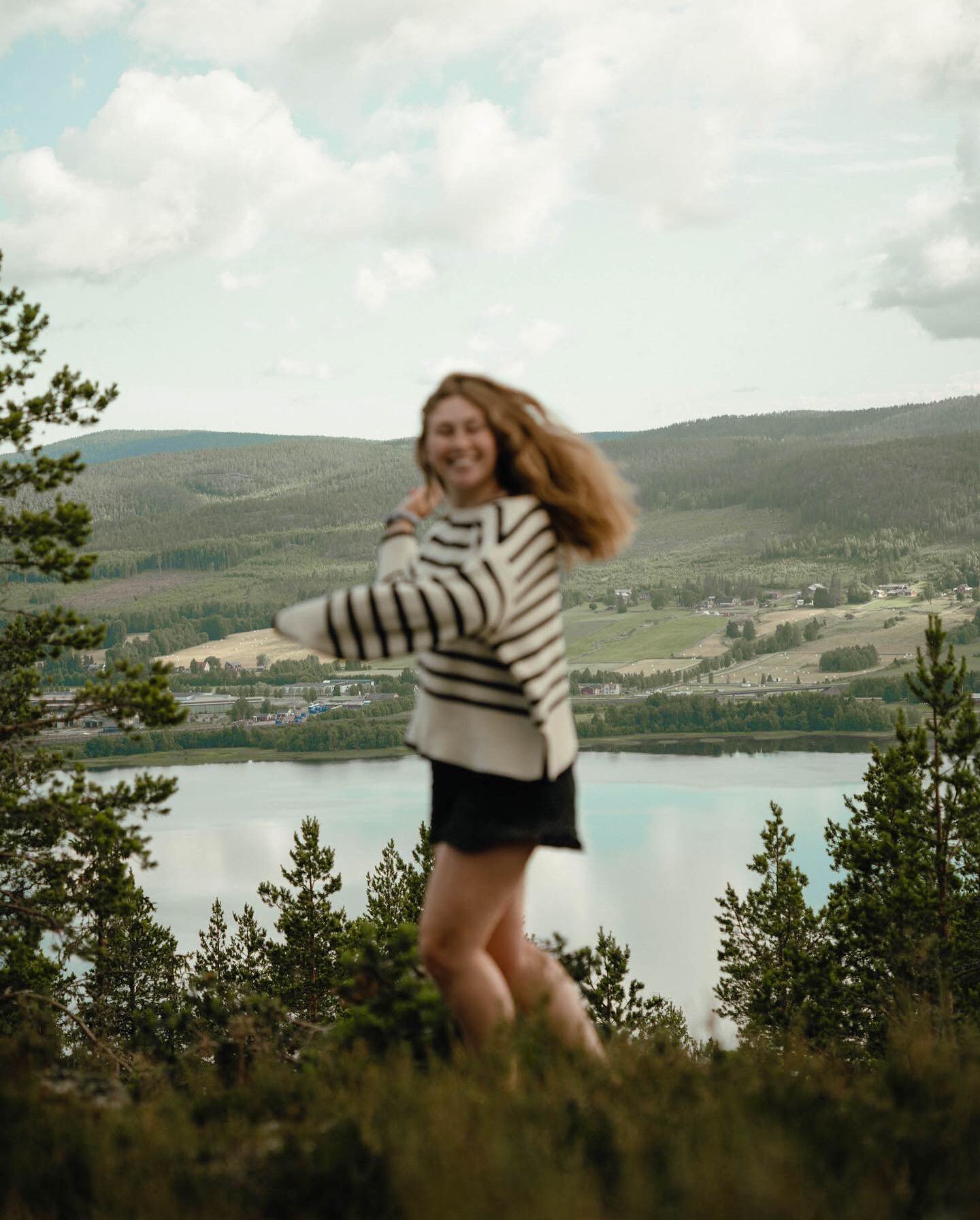 A woman with red hair in a striped sweater and black skirt standing outdoors near a lake, with trees and hills in the background, smiling and looking at the camera.
