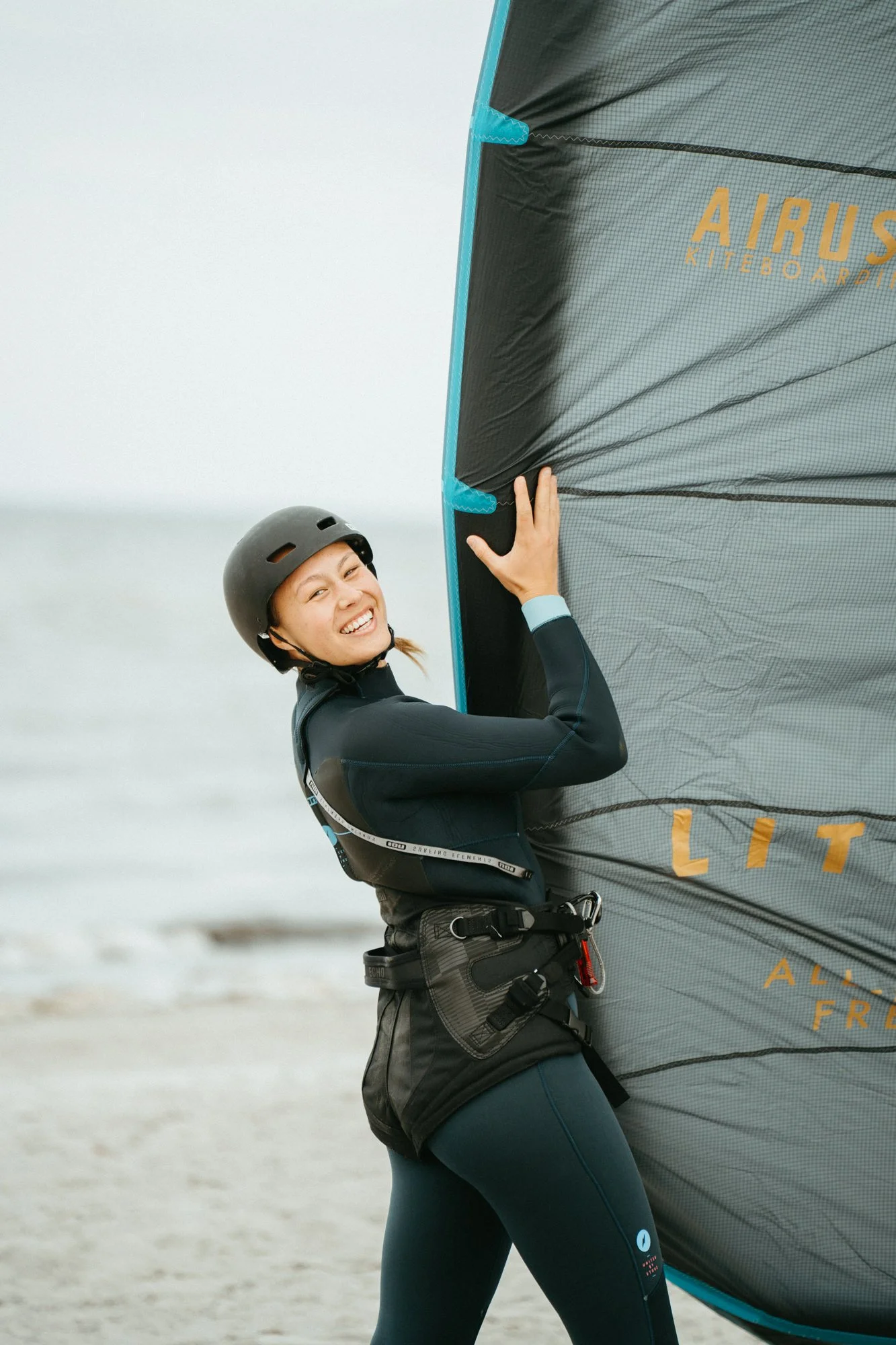 A woman smiling and wearing a helmet and wetsuit, holding a large kite by a sandy beach in front of the water.