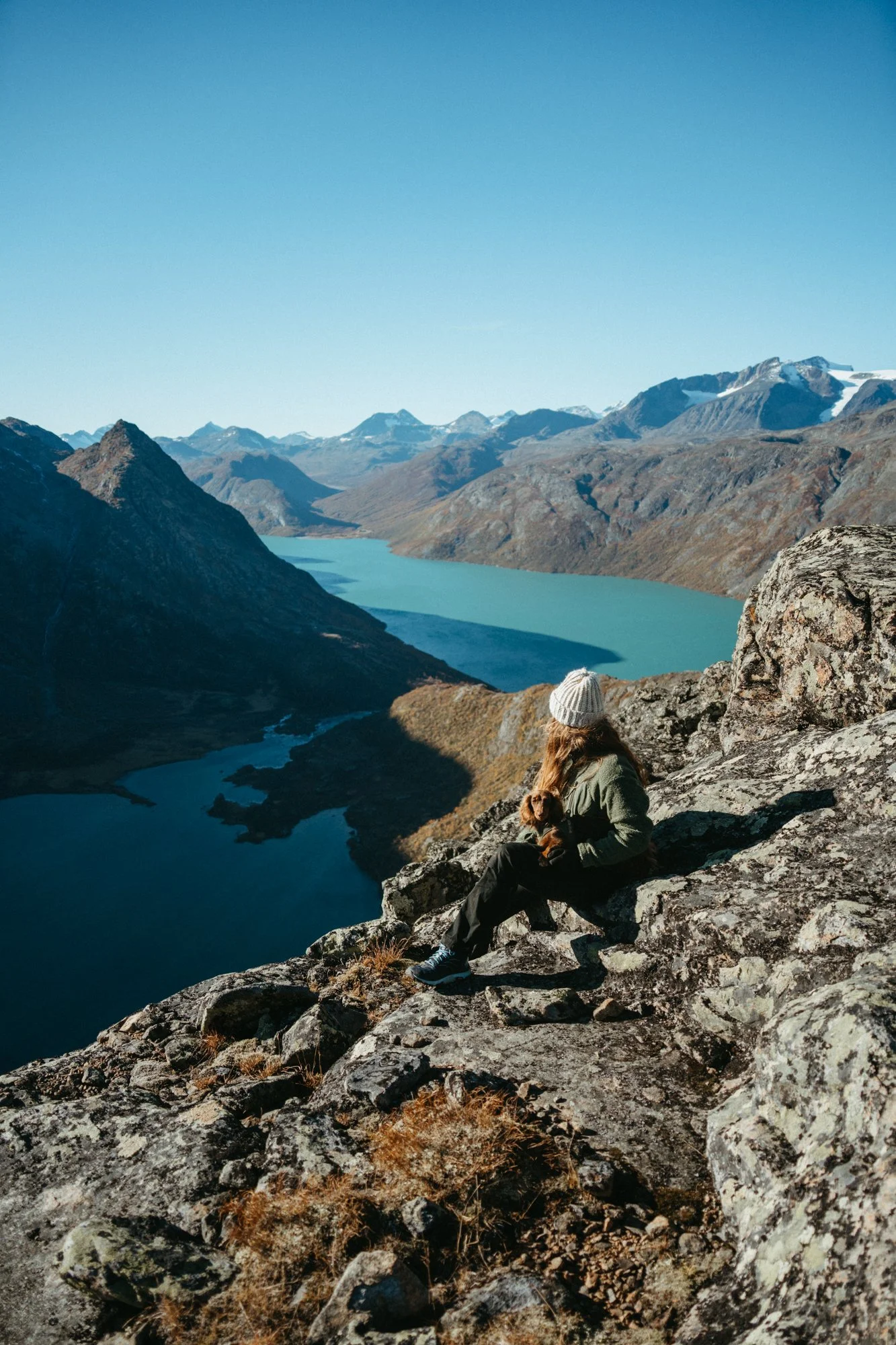 A woman sitting on rocks overlooking a lake and mountain range, holding a small dog, under clear blue sky