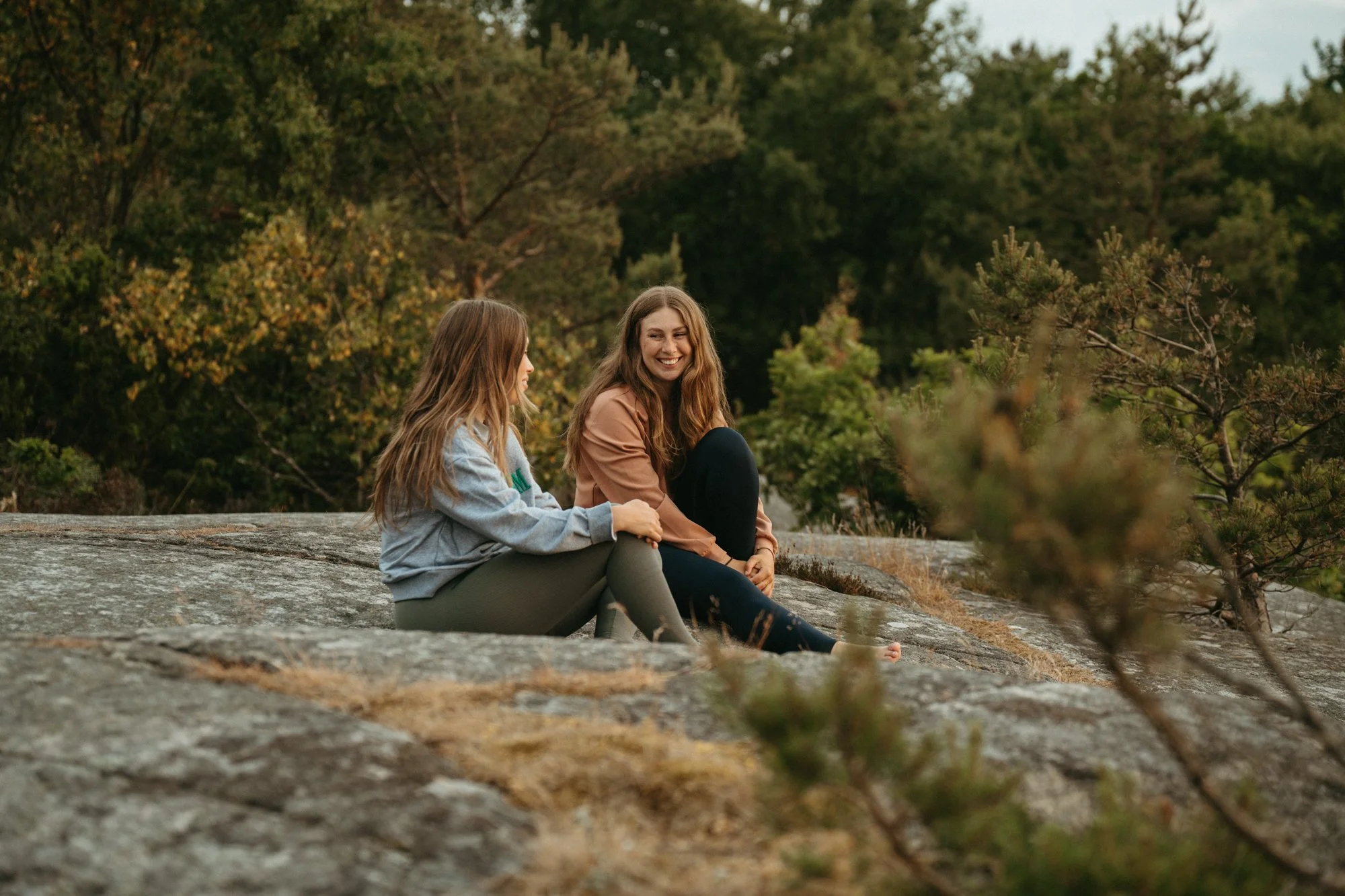 Two young women sitting on a large rock outdoors, smiling and talking, with trees and greenery in the background.