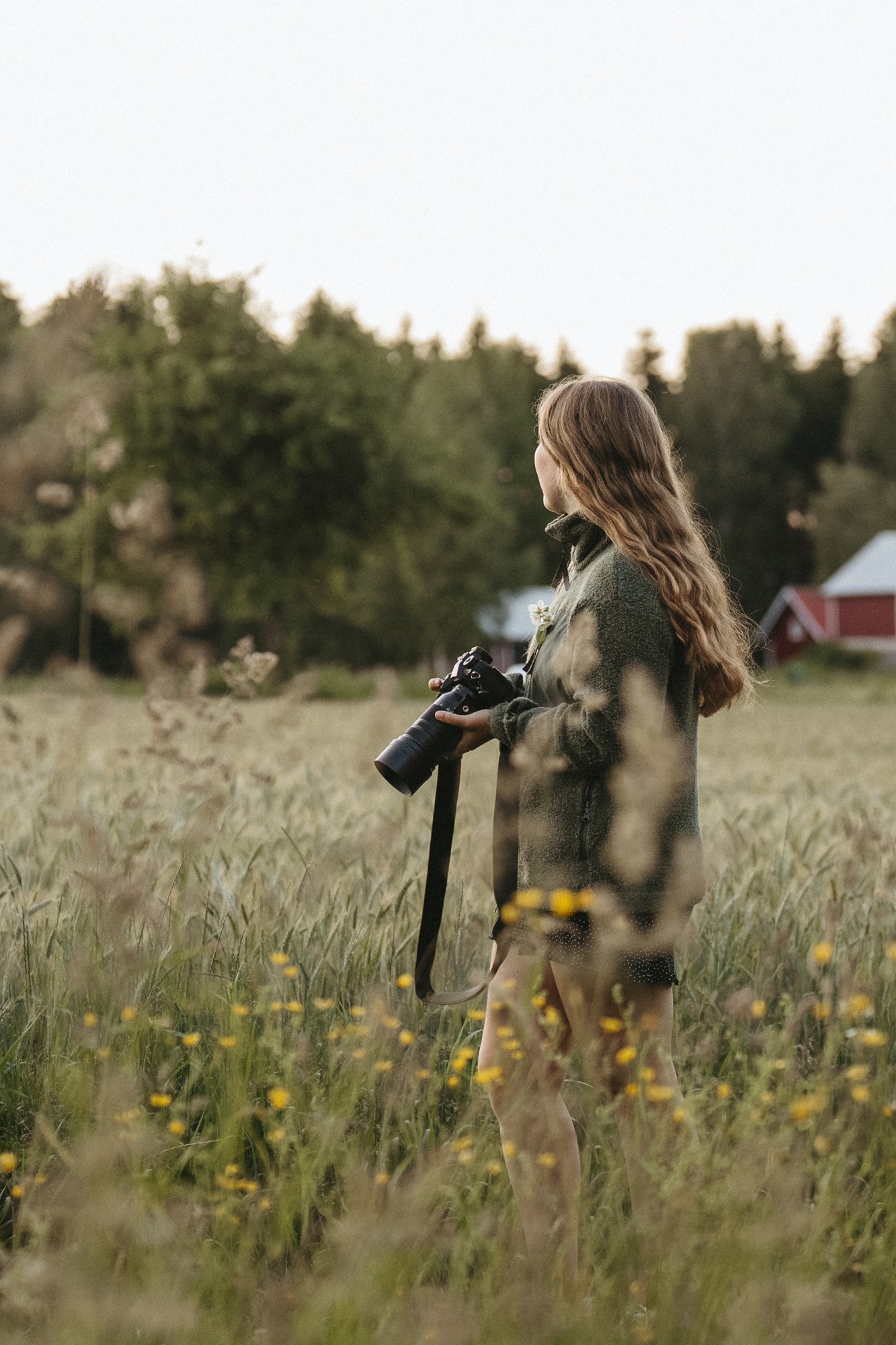 A woman with long wavy hair in a green jacket holding a camera in a field with yellow flowers, looking to the side. Trees and a red barn are visible in the background during sunset.