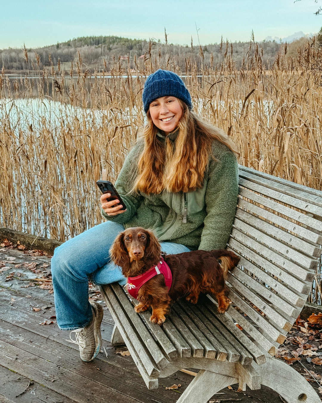A woman with long red hair wearing a blue knit hat and green fleece jacket sitting on a wooden bench with a brown dachshund in a red harness beside her near a lake and tall reeds, holding a smartphone.