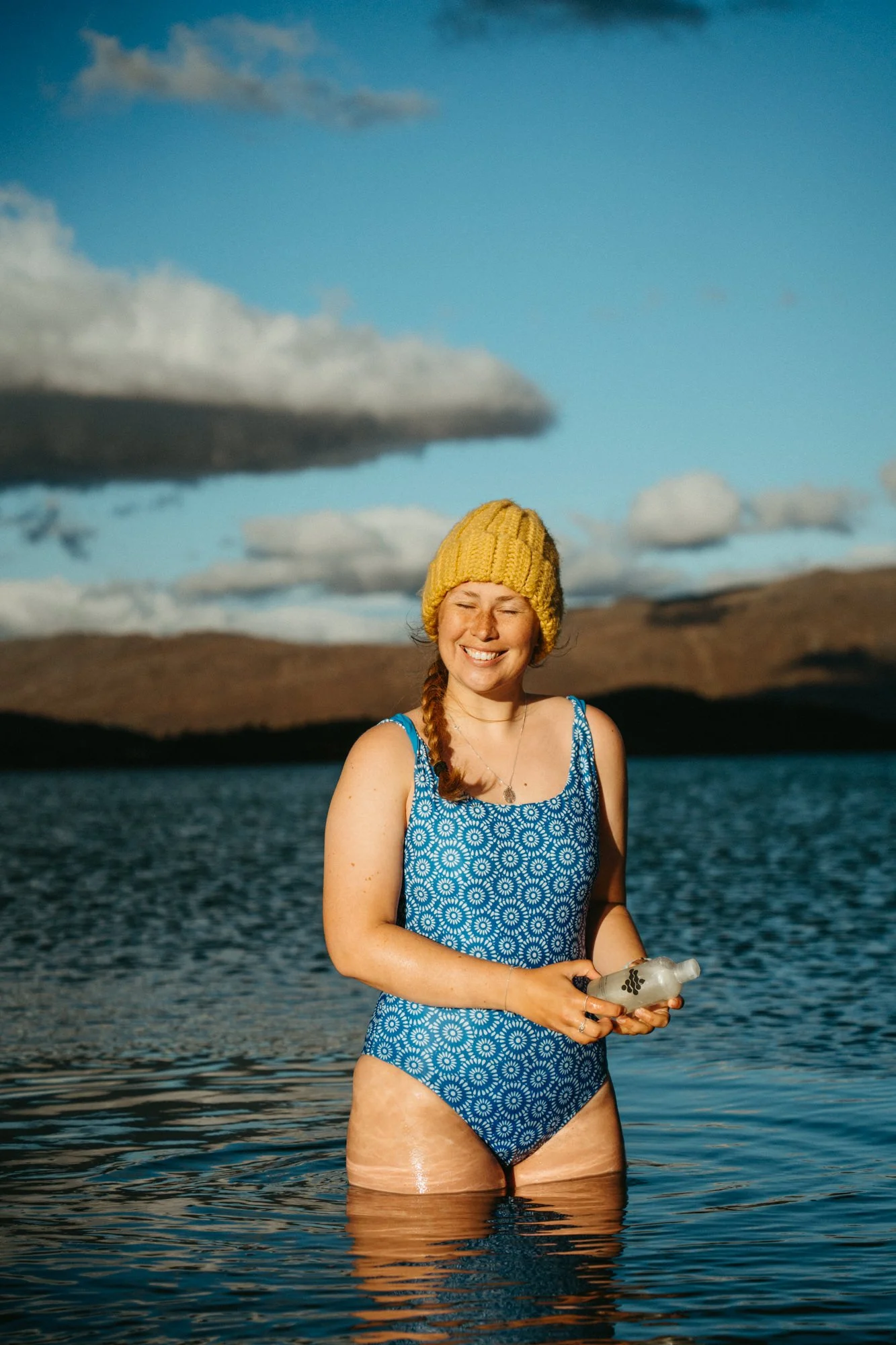 A woman wearing a blue swimsuit with white patterns, a yellow knit hat, and smiling while standing in a body of water holding a plastic bottle, with a landscape of hills and a partly cloudy sky in the background.