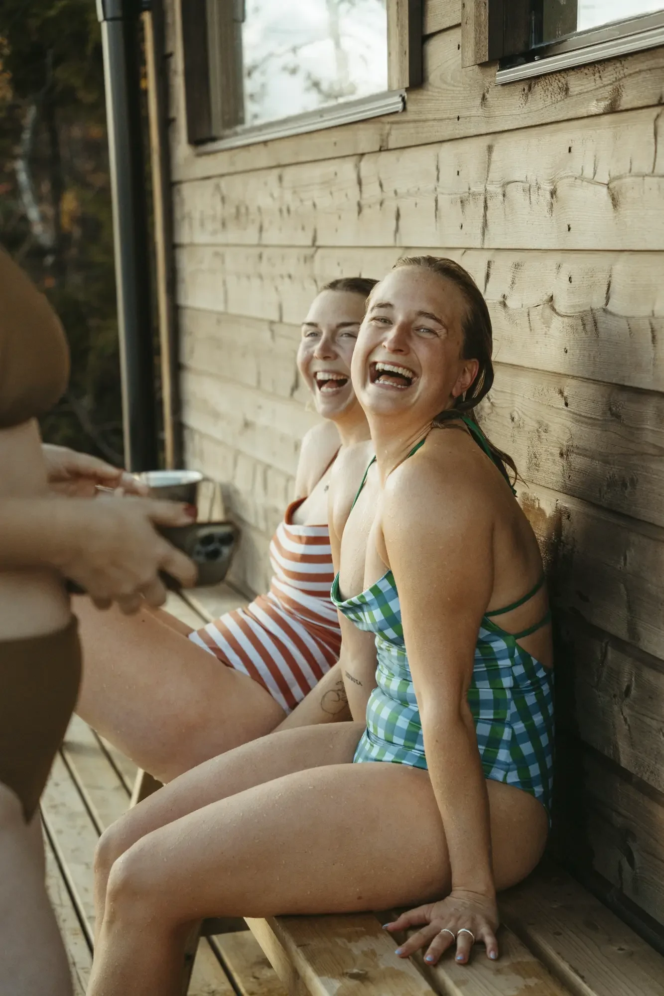 Two women in swimsuits sitting on a wooden boat or dock, smiling and laughing, with a person partially visible holding a smartphone in the foreground.