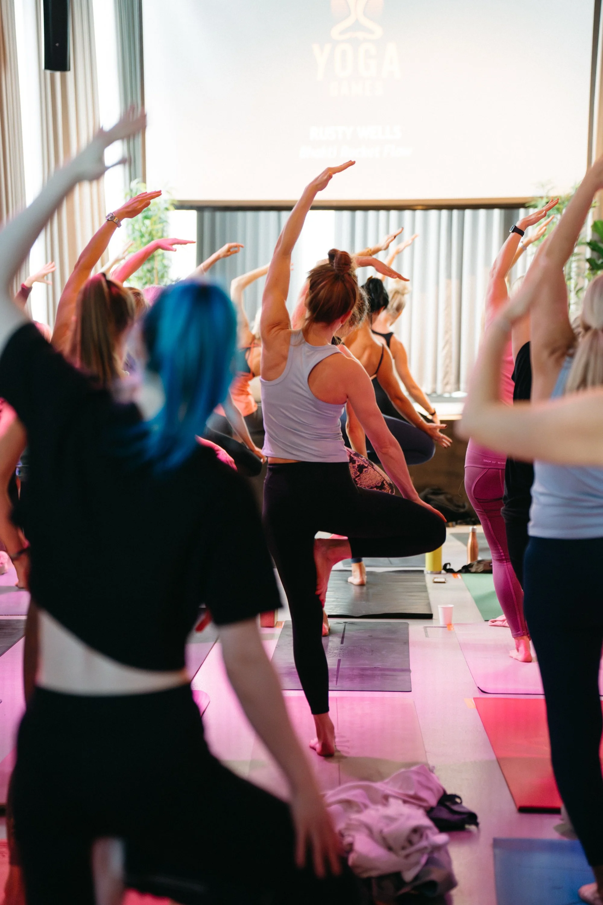 Group of women participating in a yoga class, standing on yoga mats, performing a balancing pose with one leg raised and arms extended.