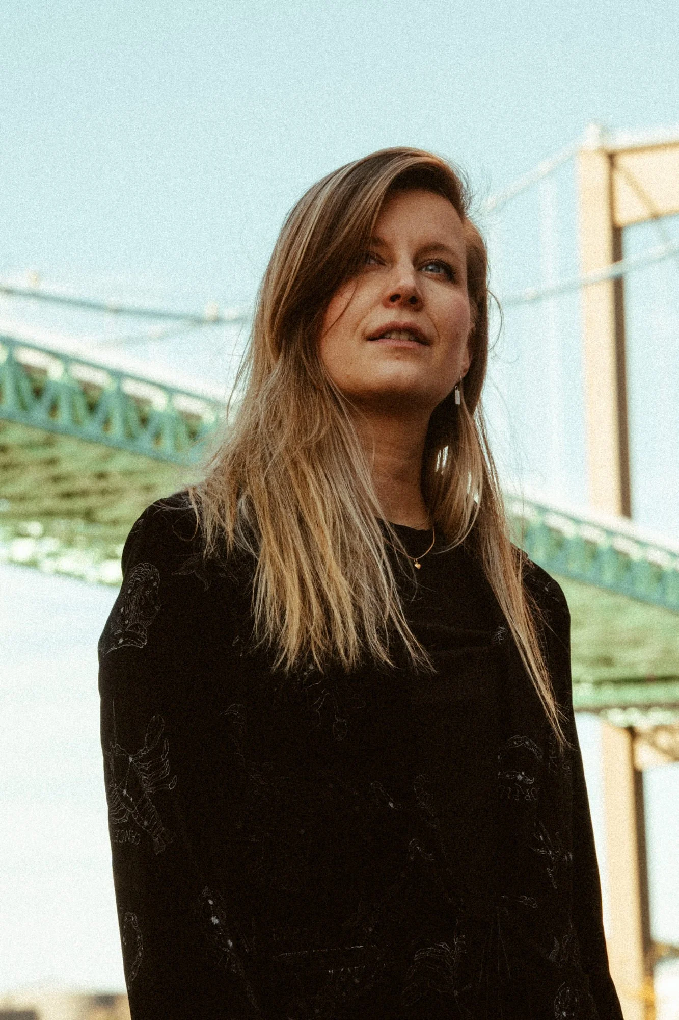 A young woman with long, wavy blonde hair stands outdoors near a bridge, looking slightly upward.