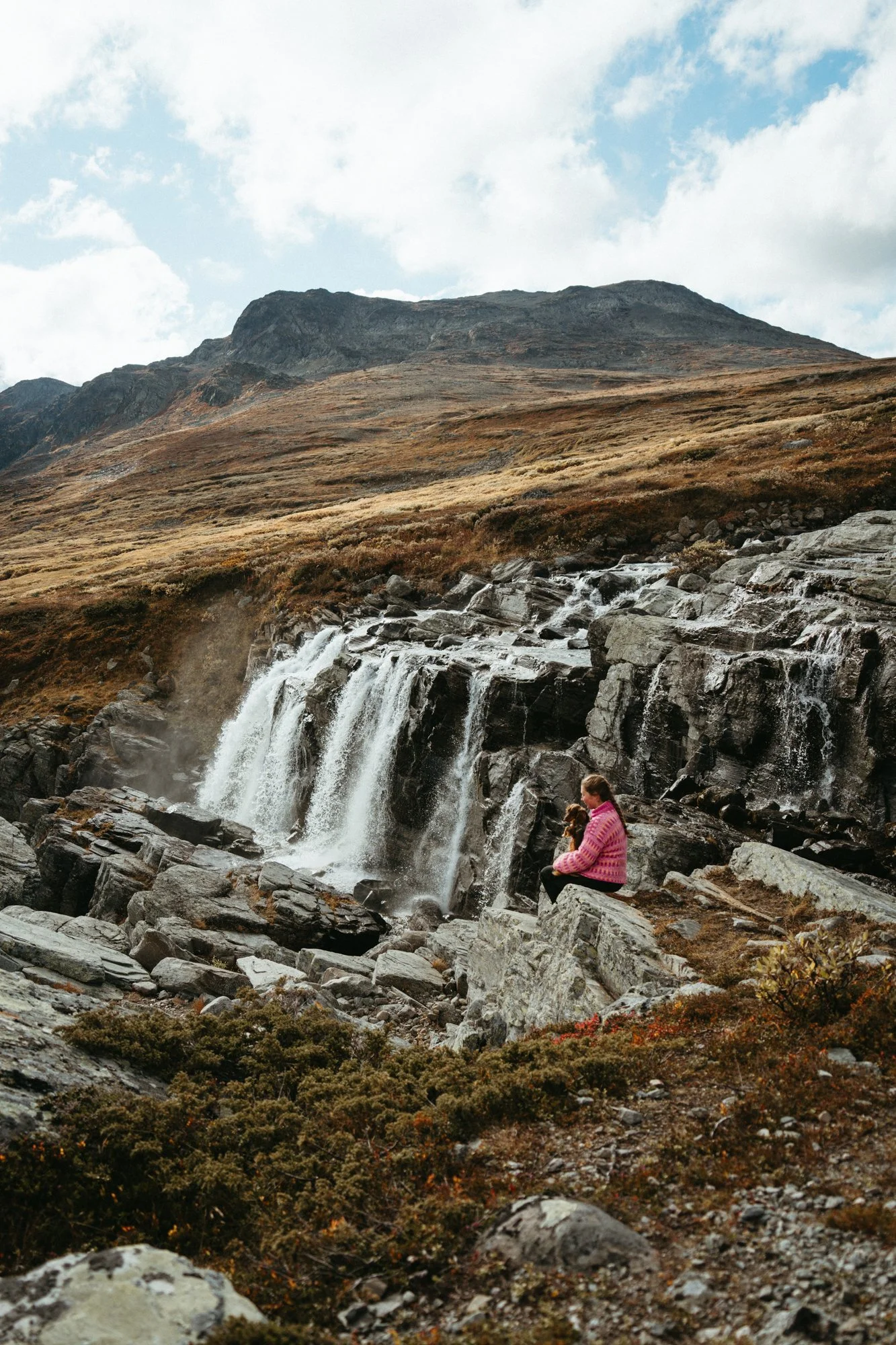 Person in pink sweater sitting on a rock near a small waterfall in a mountainous landscape.