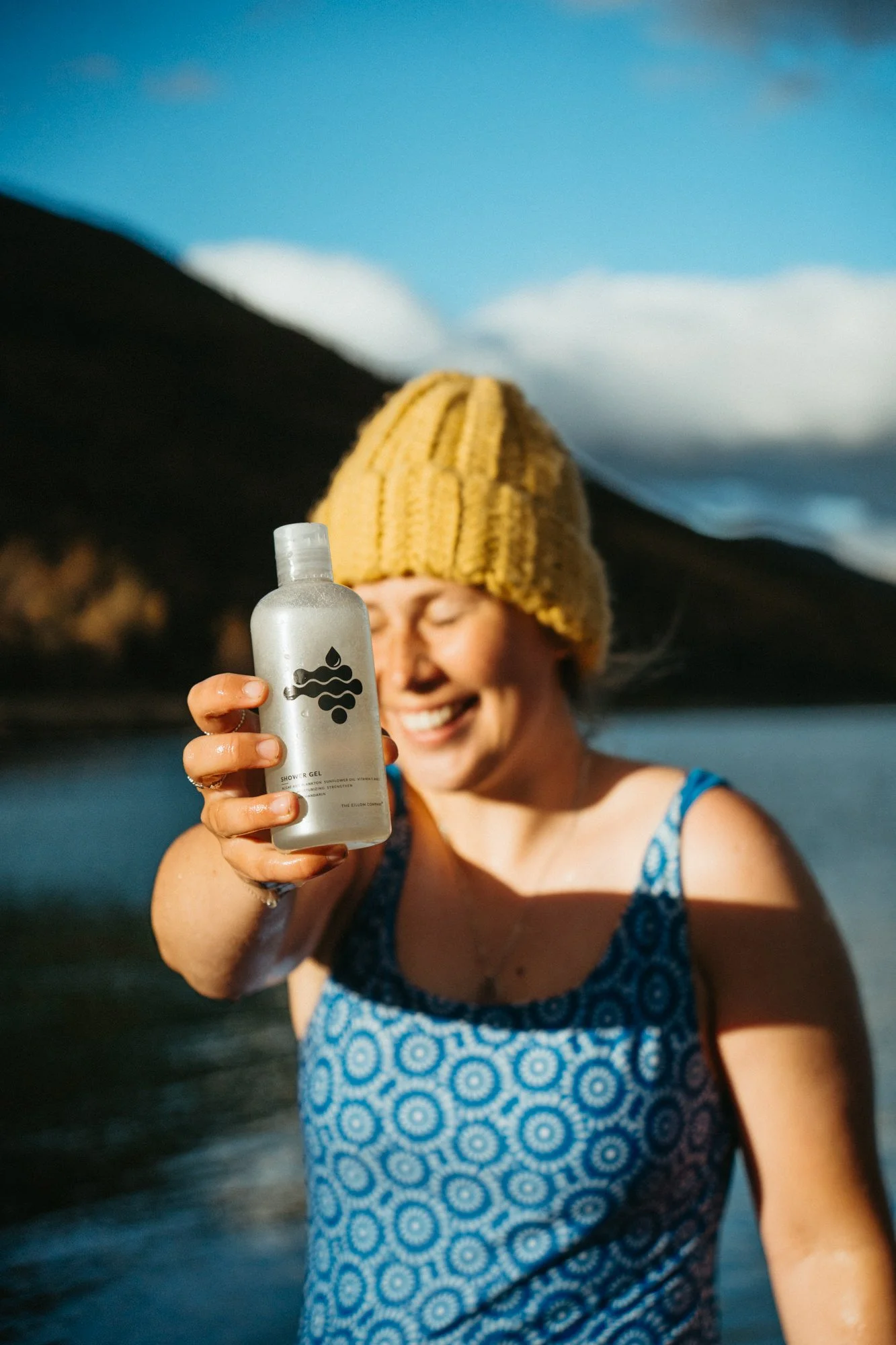 Woman in blue patterned dress and yellow knit hat holding up a silver bottle of shower gel with a water and wave design on it, smiling outdoors near a body of water with hills and a blue sky in the background.