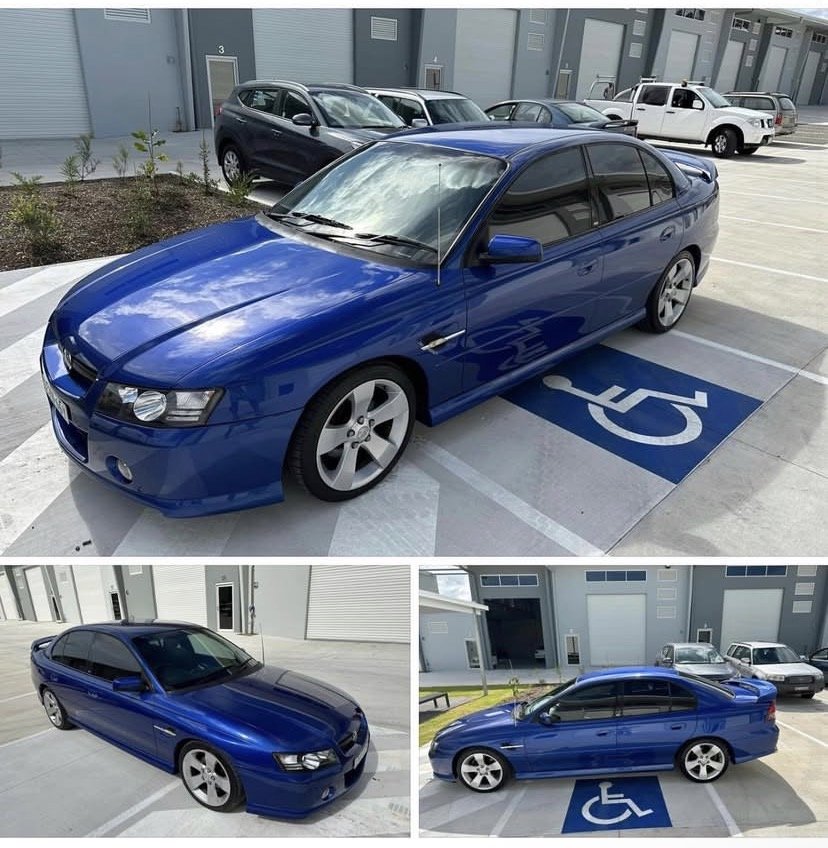 Blue sedan parked in a designated handicapped parking space with a large handicapped symbol painted on the ground, and other parked cars and a building in the background.