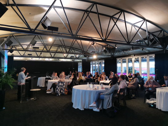 A group of people attending a presentation or workshop in a modern, spacious conference room with large windows, tablecloth-covered tables, and a presenter standing at a podium.