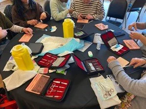 People sitting around a table with makeup kits, bottles, tissues, and smartphones during a makeup session or workshop.