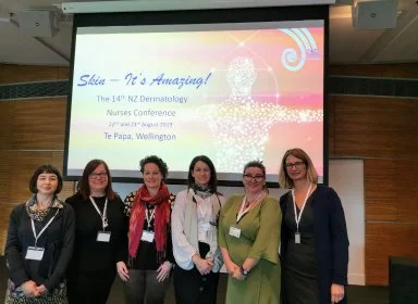 Group of six women standing in front of a presentation screen at a conference, smiling and wearing conference badges.