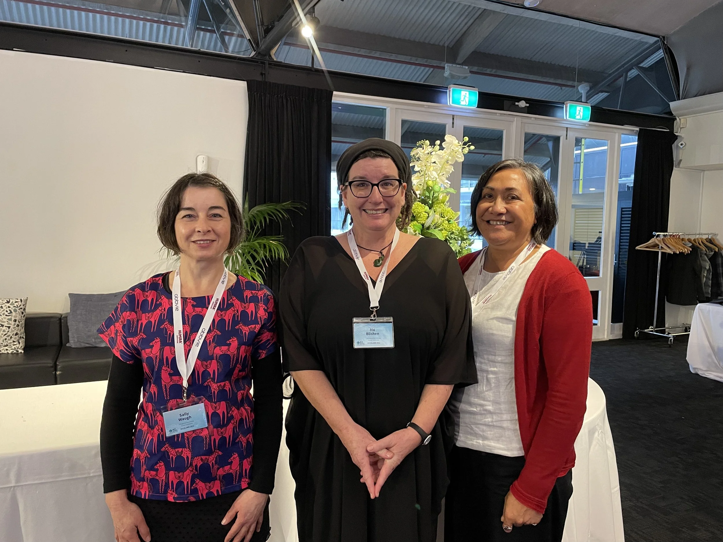 Three women standing together indoors, smiling for the camera. The woman on the left wears a red and blue patterned shirt and a white lanyard. The woman in the middle wears black glasses and a black dress, with a plant and window behind her. The woman on the right wears a white shirt and red cardigan, with a rack of clothing in the background.