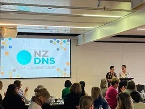 A presentation in a conference room with a large screen displaying the NZ DNS logo and people sitting at tables