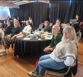 People seated at round tables during a conference or meeting in a dimly lit room.