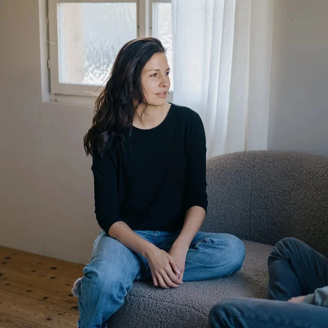 Eine Frau sitzt entspannt auf einer Couch in einem Raum mit natürlichem Licht aus einem Fenster.