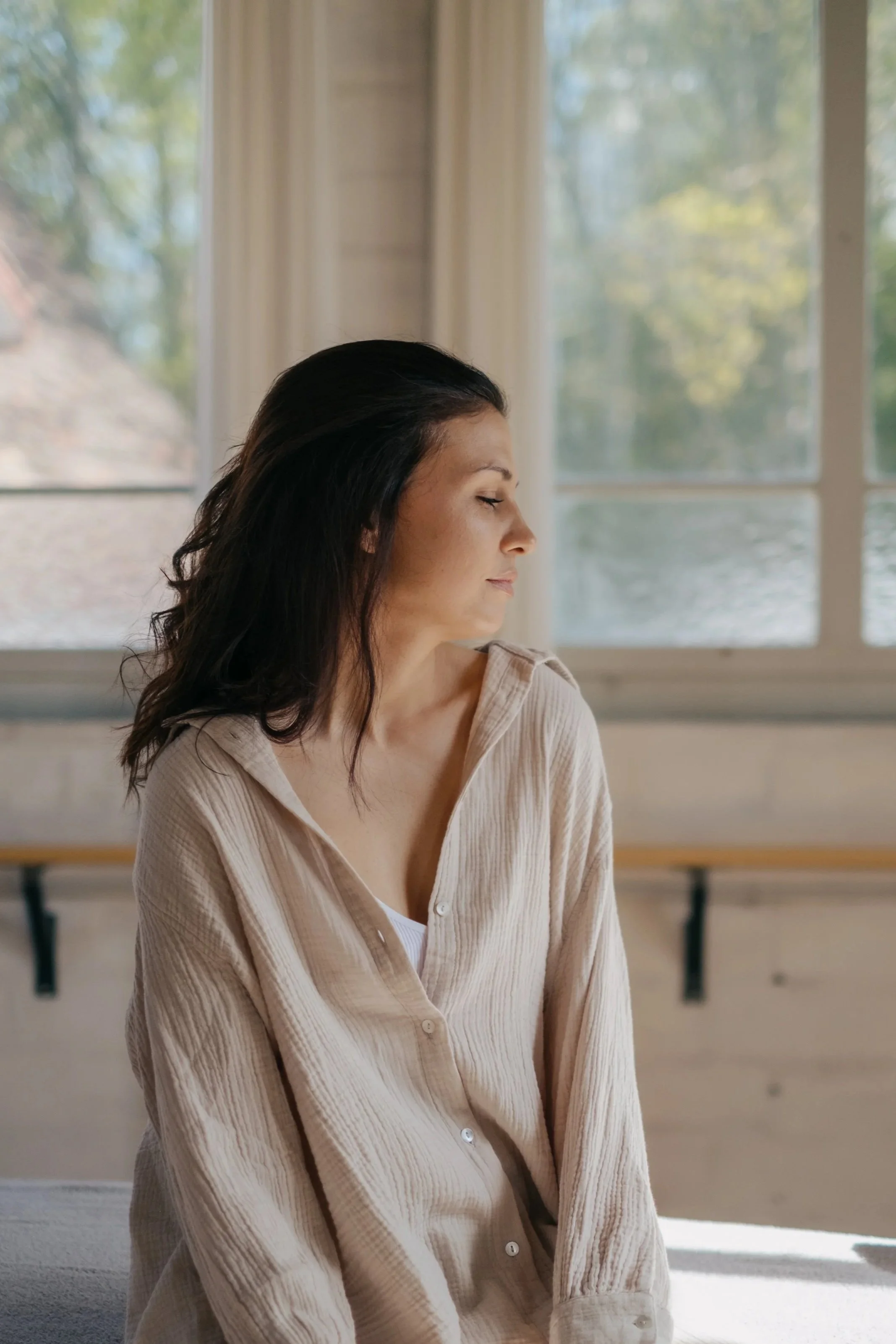 Eine Frau sitzt mit geschlossenen Augen auf einer Massageliege in einem hellen Raum vor einem Fenster.