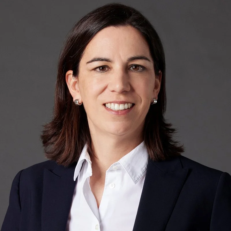 Professional headshot of a woman with dark brown hair, wearing a white collared shirt and a black blazer, smiling against a gray background.