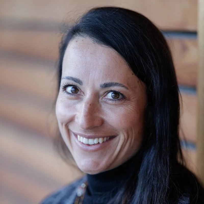 A woman with dark hair smiling in front of a wooden wall.