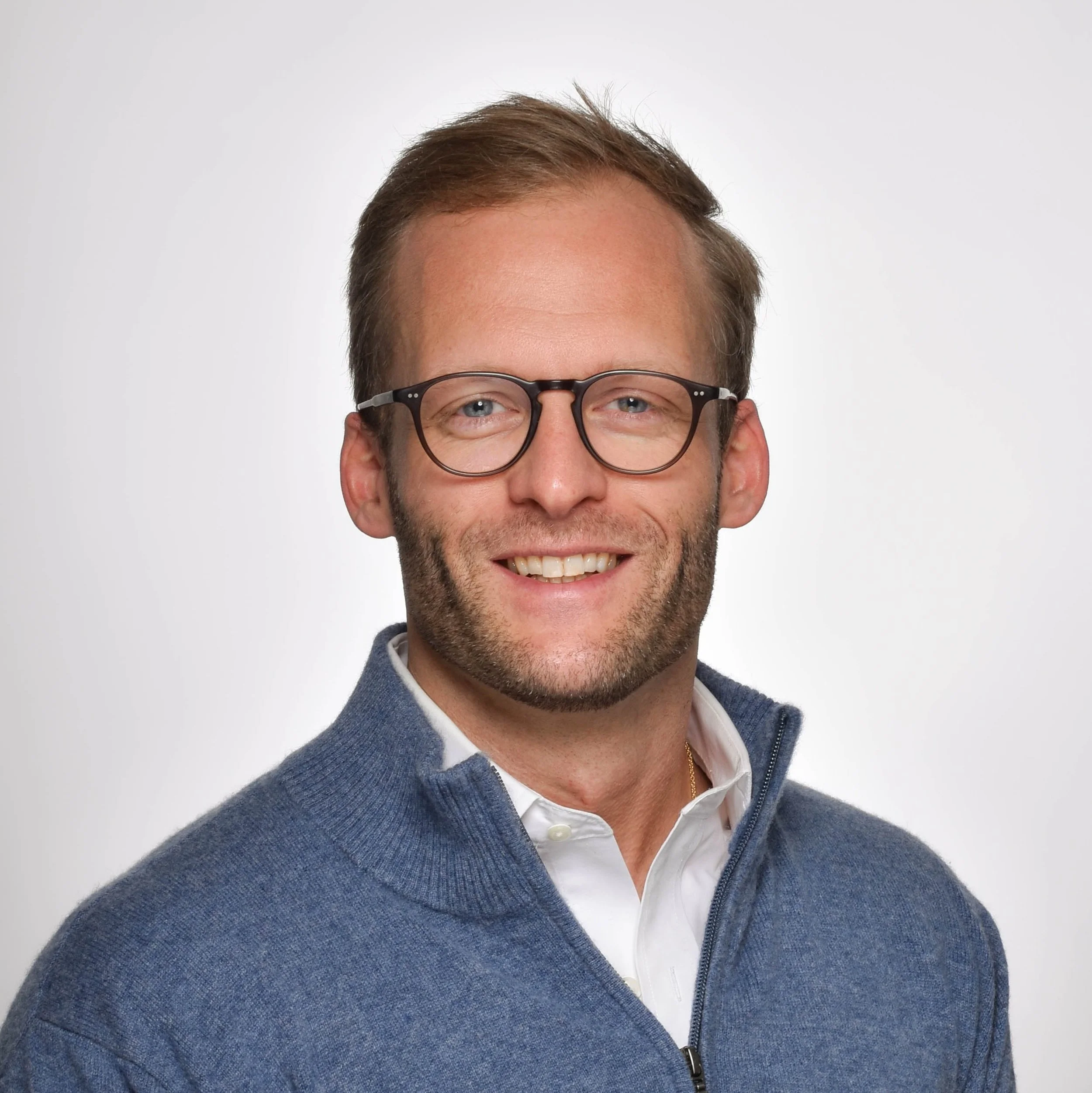 Portrait of a smiling man with glasses, wearing a white shirt and a blue zip-up sweater, against a plain white background.
