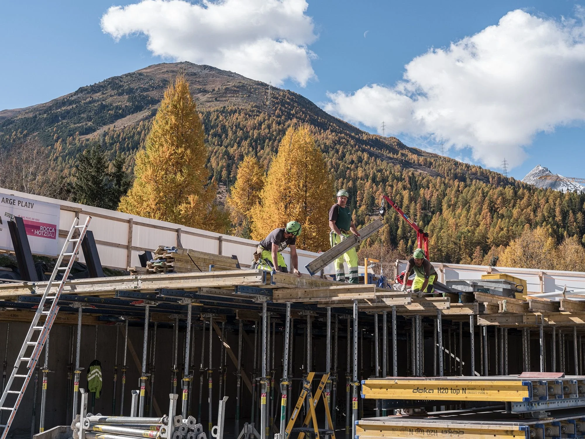Construction workers building a structure with wooden beams on scaffolding, with mountains and trees in the background.