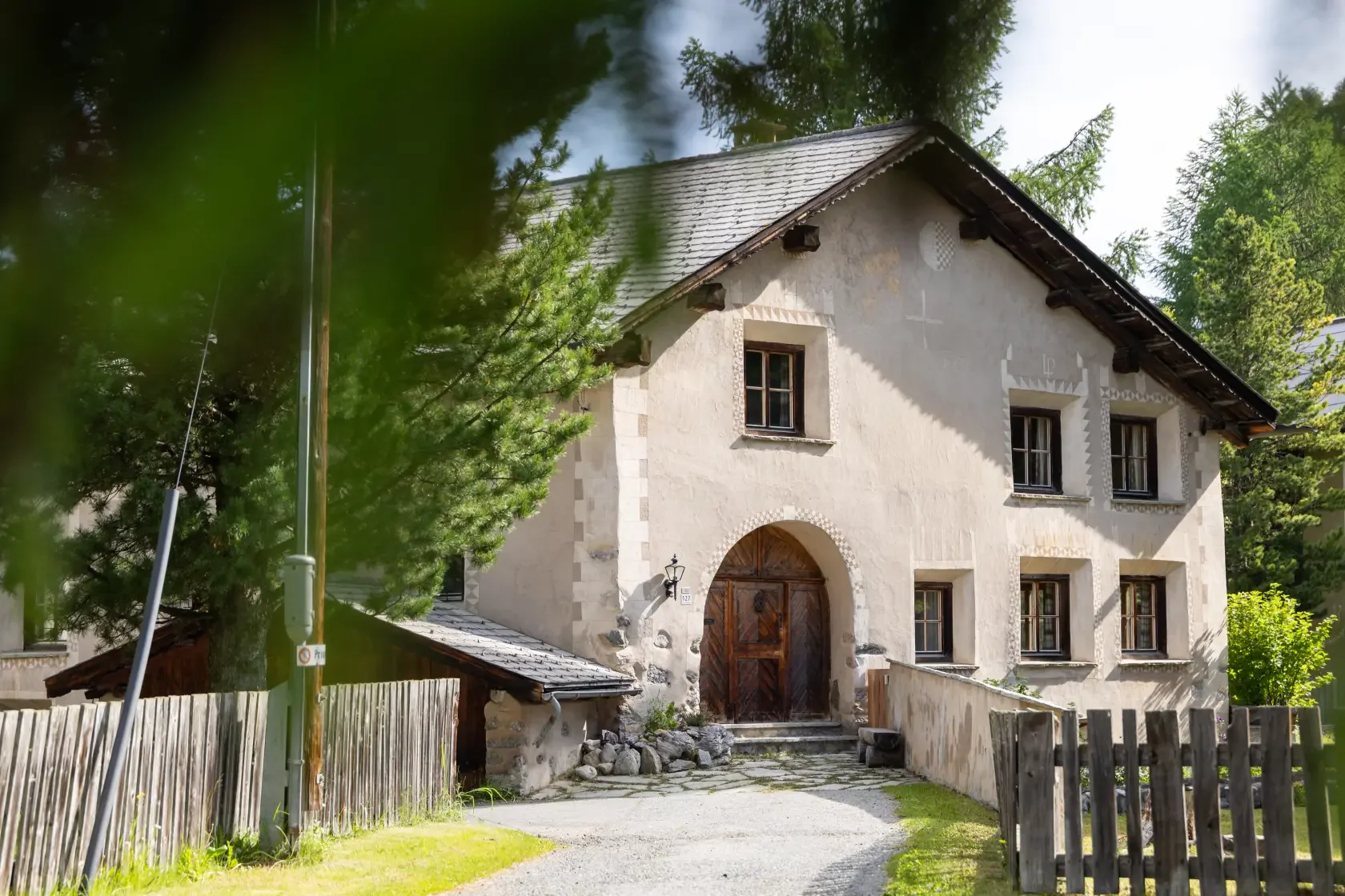 A rustic two-story house with a wooden arched front door, surrounded by green trees and a wooden fence, with a gravel pathway leading to the entrance.