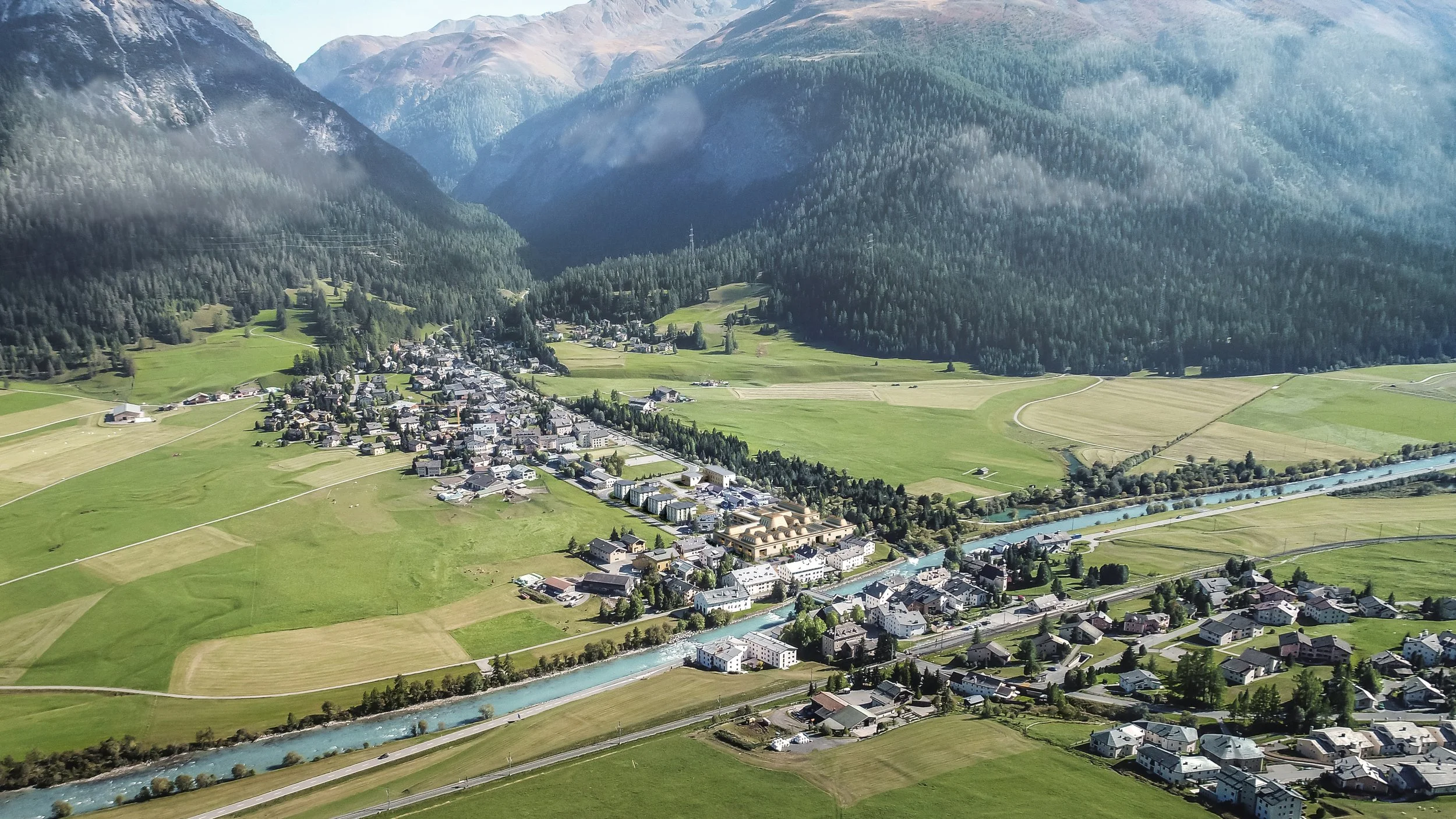 Aerial view of a mountain town with a river running through it, surrounded by green fields and forested mountains.