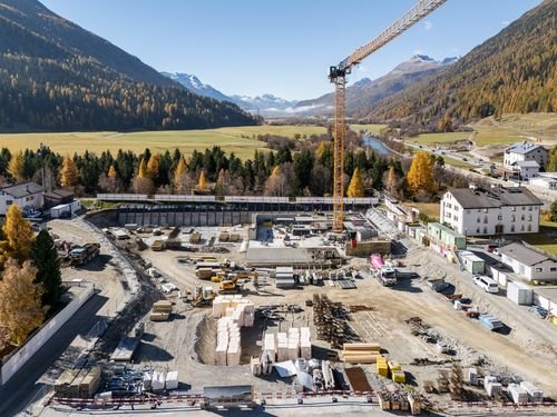 Construction site in a valley surrounded by mountains, with a crane, building materials, and partially built structures.