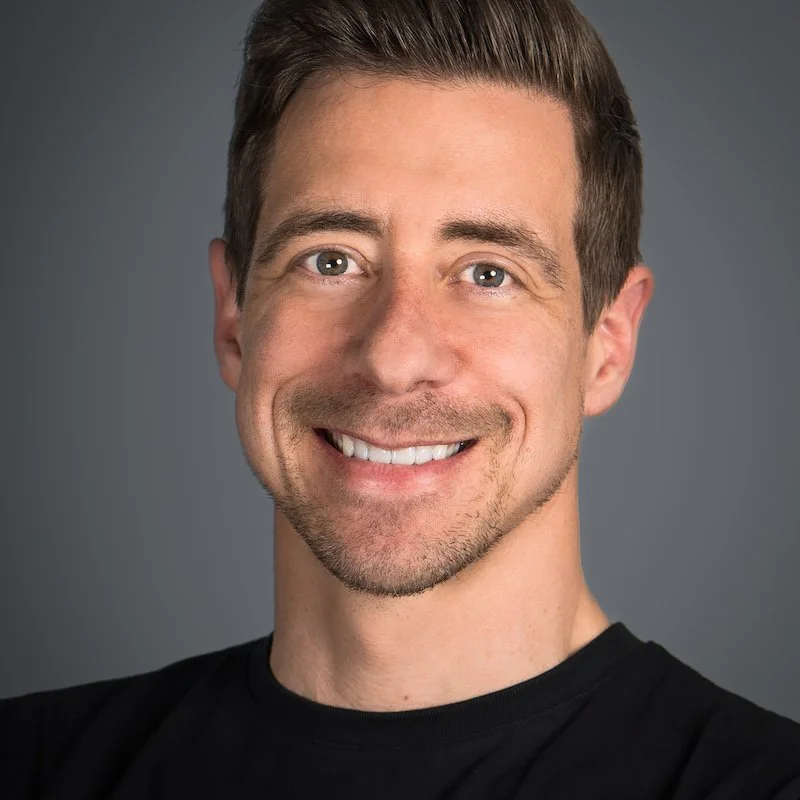 Smiling man with brown hair, light blue eyes, and light stubble, wearing a black shirt against a gray background.