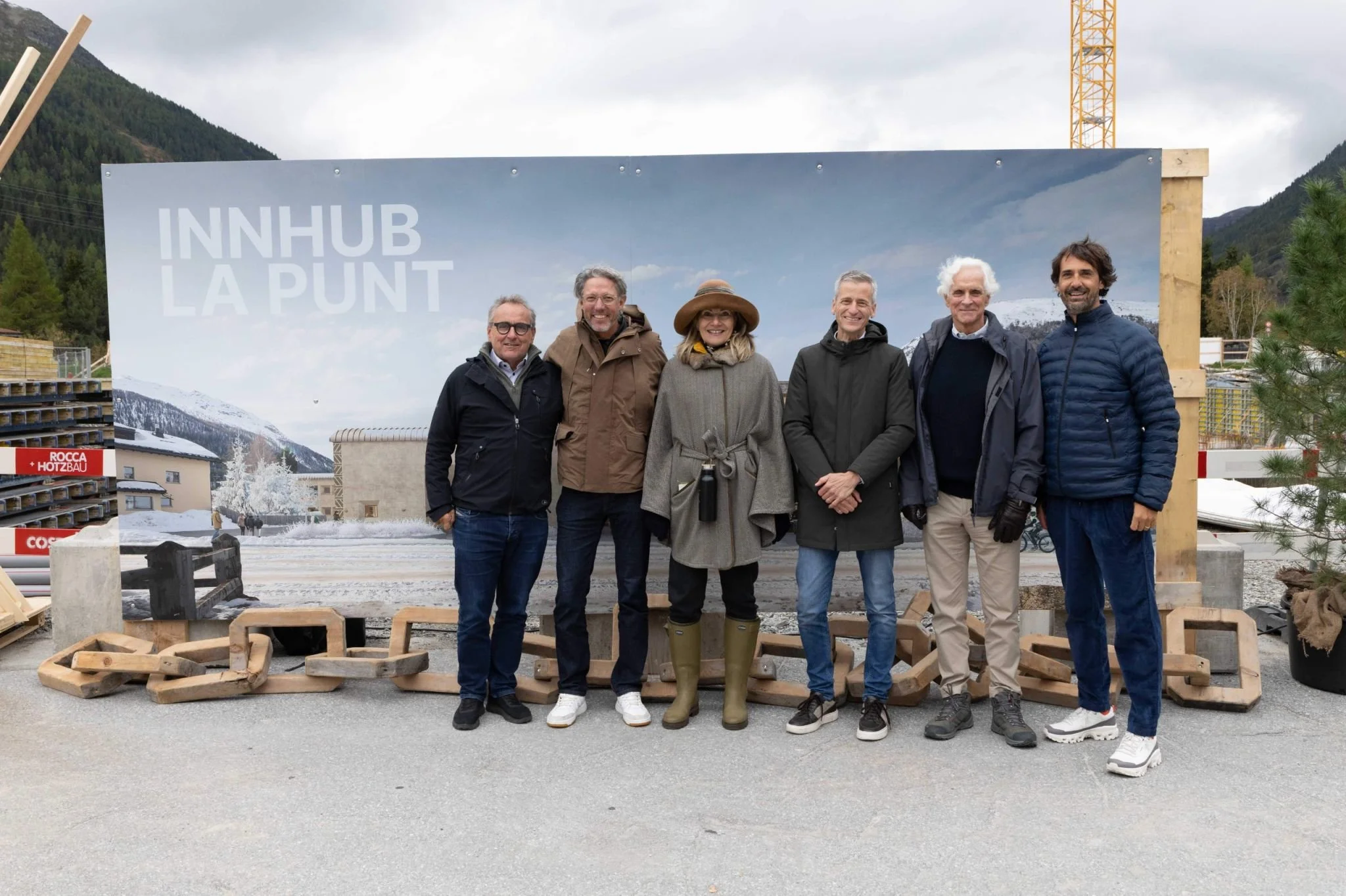 Group of six people standing outdoors in front of a large sign that reads 'INNHUB LA PUNT'. The background includes mountainous terrain and cloudy sky, with some snow on the ground and mountains. The individuals are dressed in jackets and casual clothing, posing for the photo.
