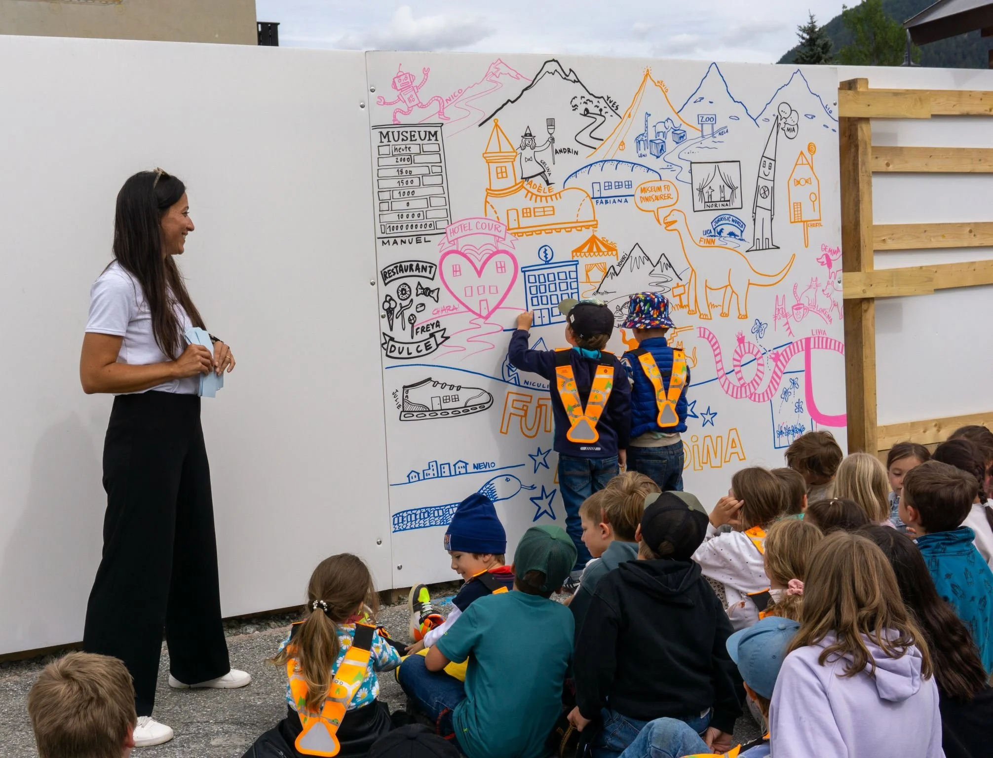 A woman and two children drawing on a large illustrated outdoor mural, surrounded by seated children.