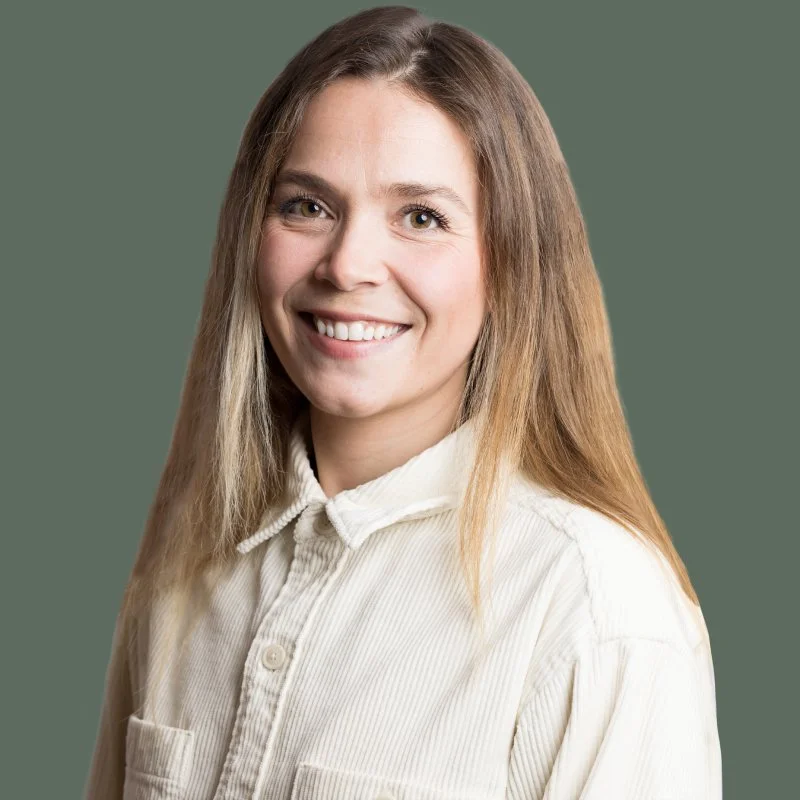 A young woman with long light brown hair and blue eyes smiling at the camera, wearing a white button-up shirt, against a green background.
