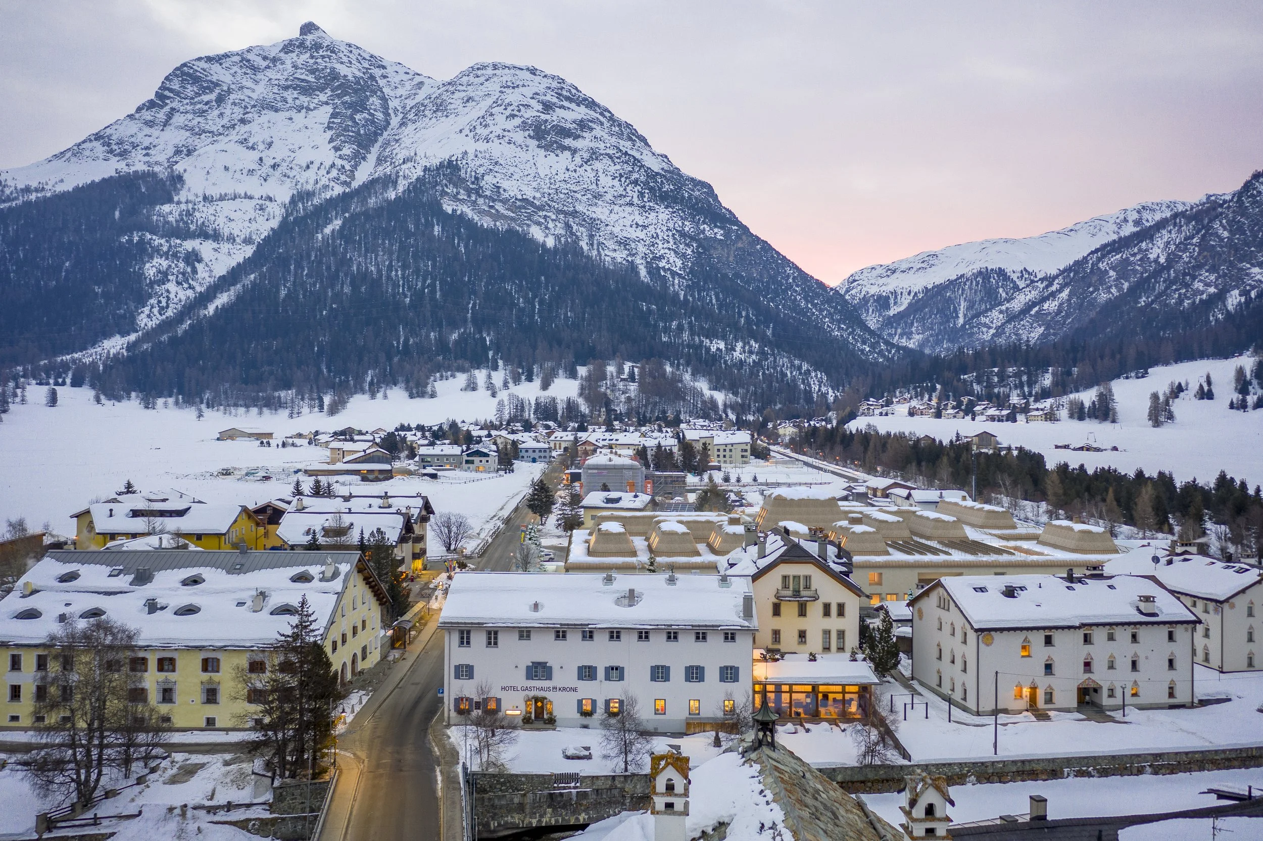 Snow-covered village with yellow, white, and beige buildings in a valley surrounded by tall, snow-capped mountains and evergreen trees.
