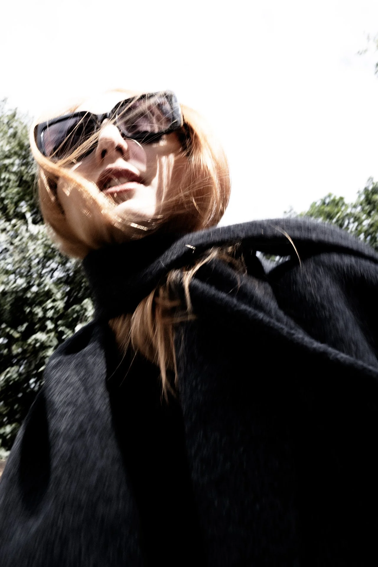 Low-angle photo of a woman with red hair wearing sunglasses and a black coat, outdoors on a sunny day with trees in the background.