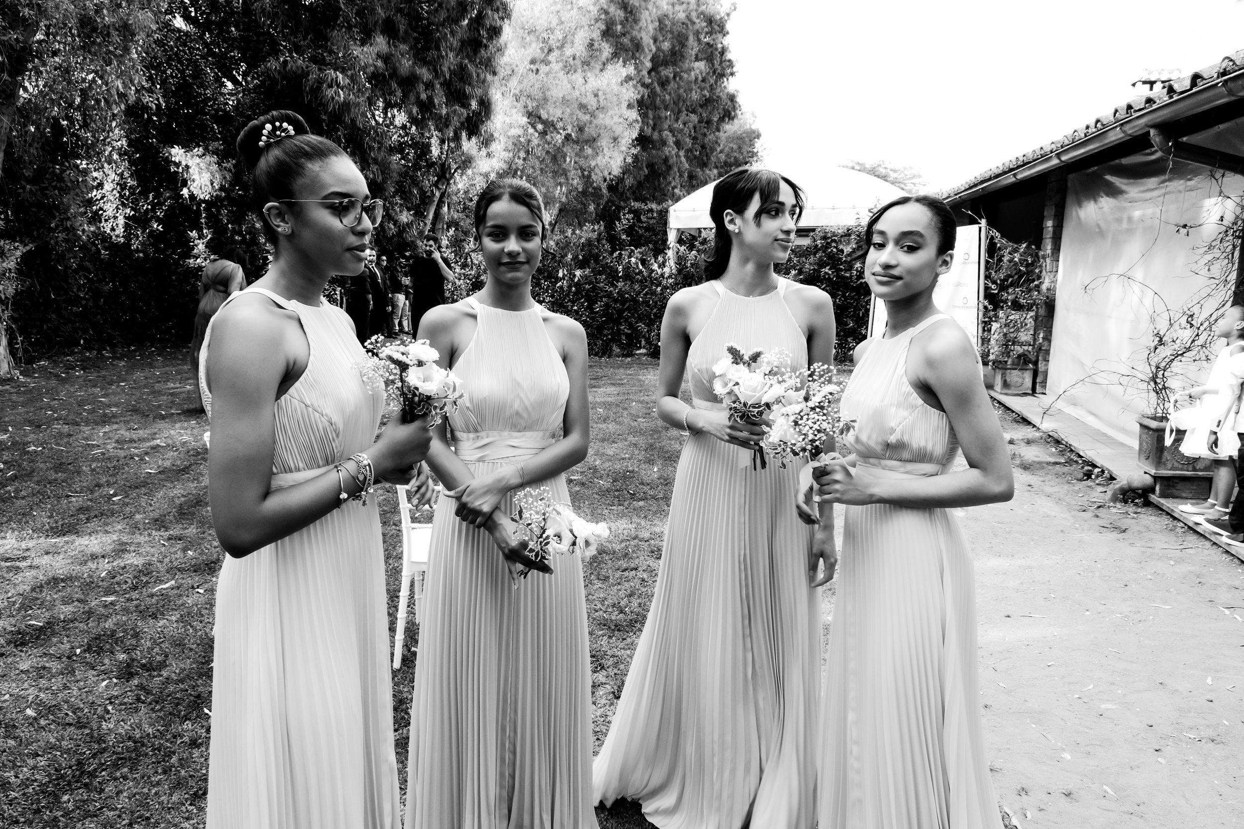 Four women in long, light-colored dresses holding bouquets on a garden or outdoor venue.